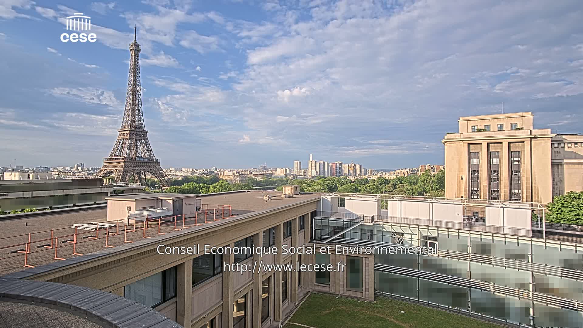 A rooftop view of Paris on a partly cloudy day shows the Eiffel Tower in the distance, with other buildings and greenery in the foreground.