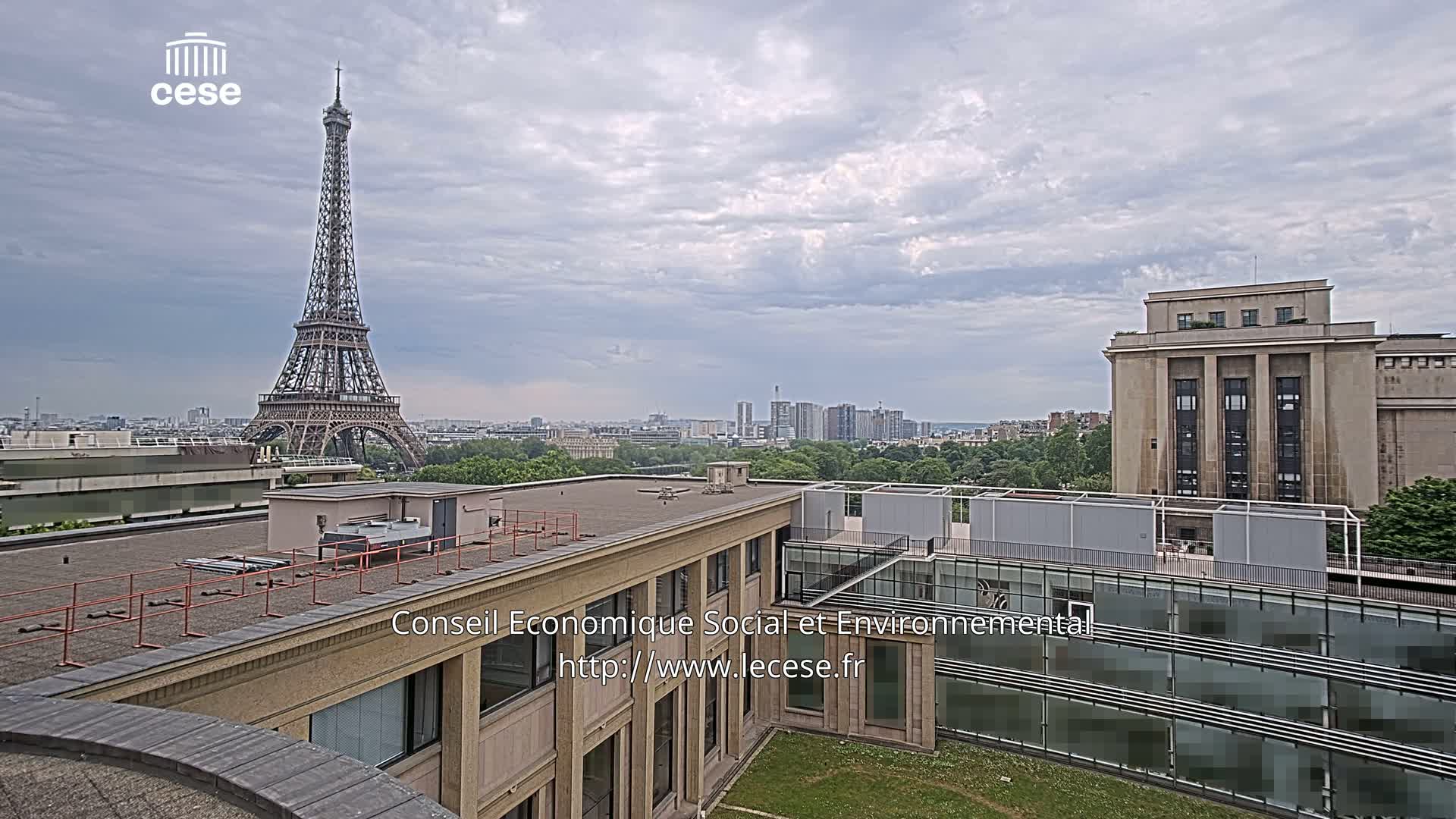A rooftop view shows the Eiffel Tower in the distance under a cloudy sky, with a city skyline and other buildings visible.