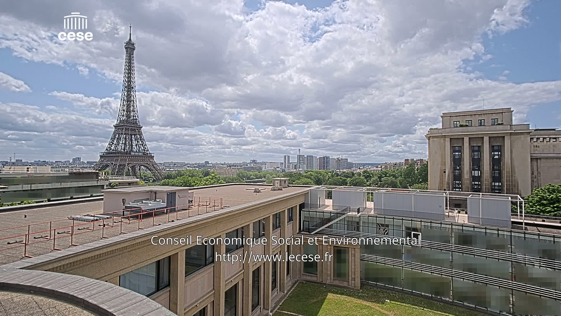 A view from a rooftop shows the Eiffel Tower in the distance under a partly cloudy sky, with other buildings and greenery in the foreground.