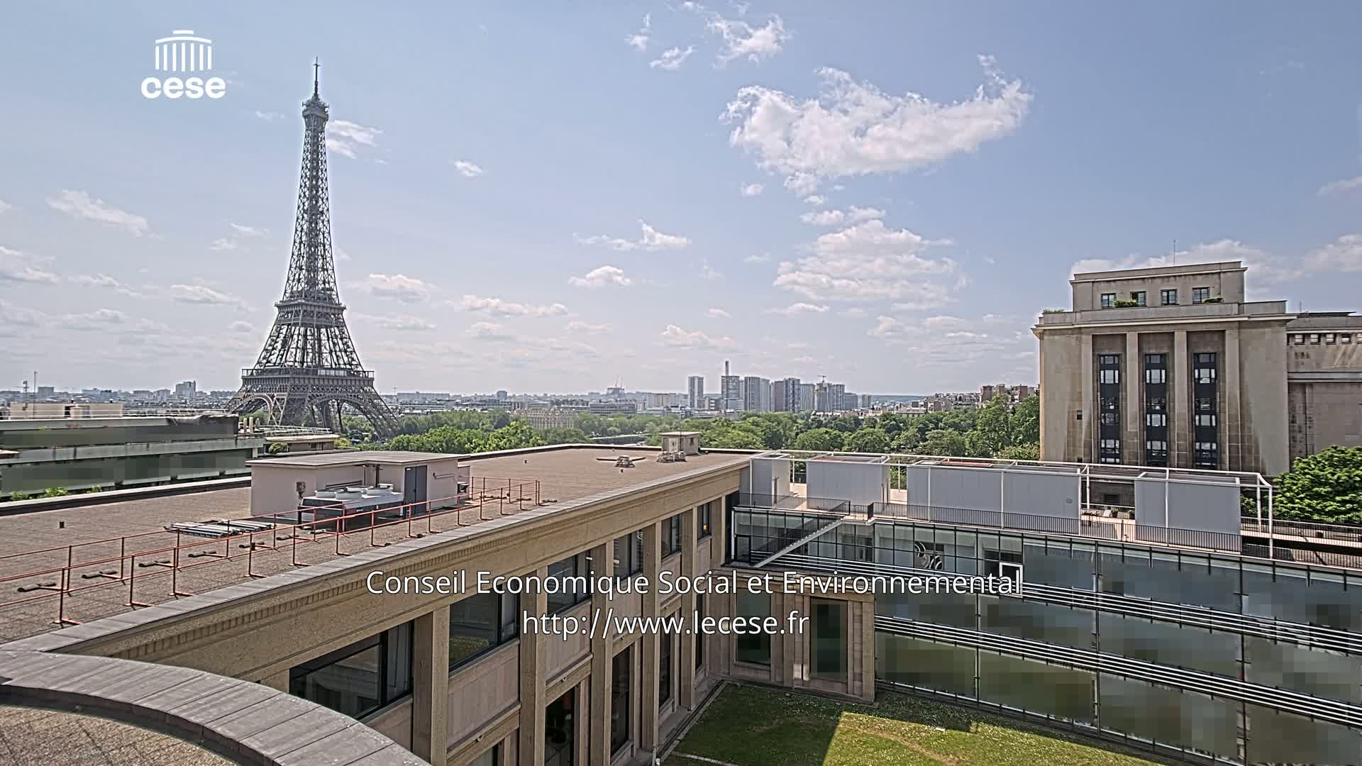 A rooftop view of Paris on a partly cloudy day shows the Eiffel Tower in the distance, along with other buildings and green spaces.