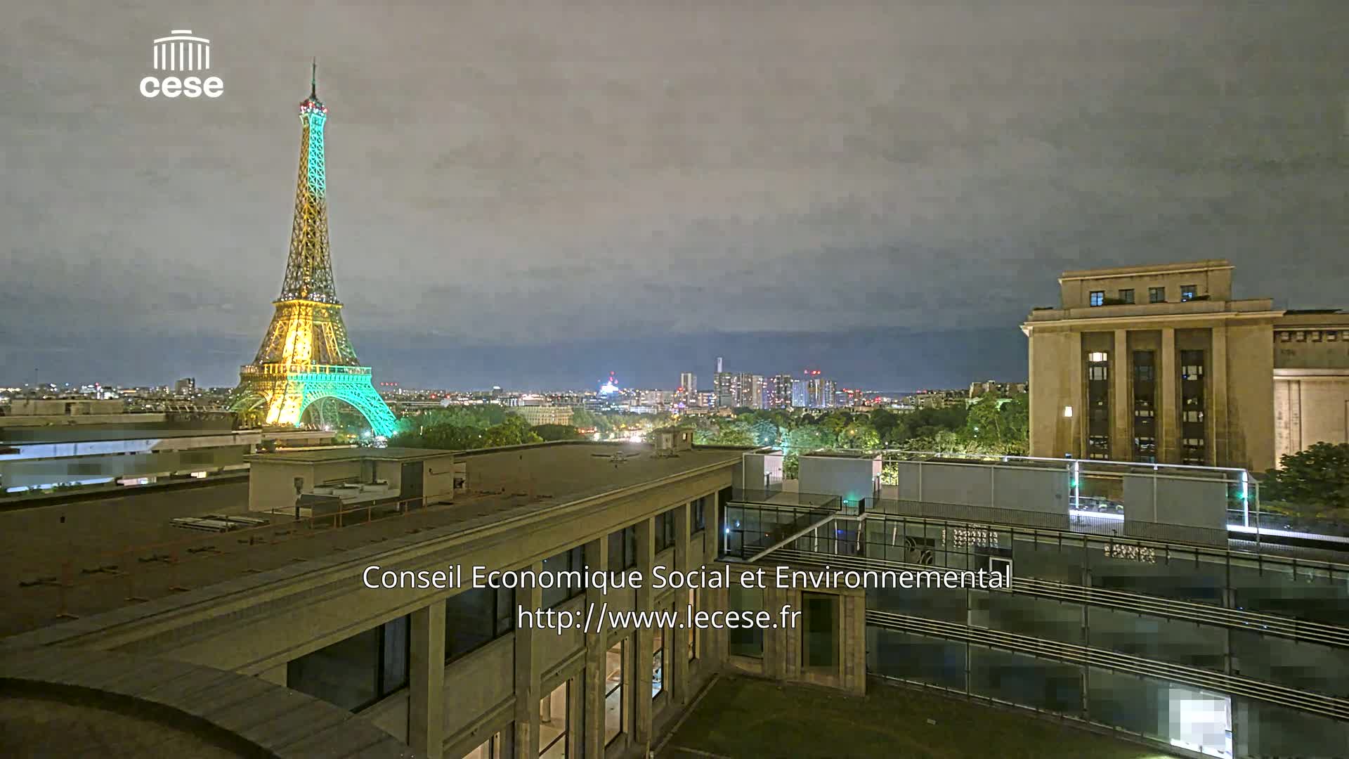 A nighttime view of the Eiffel Tower illuminated in teal and gold, alongside a cityscape under a cloudy sky.