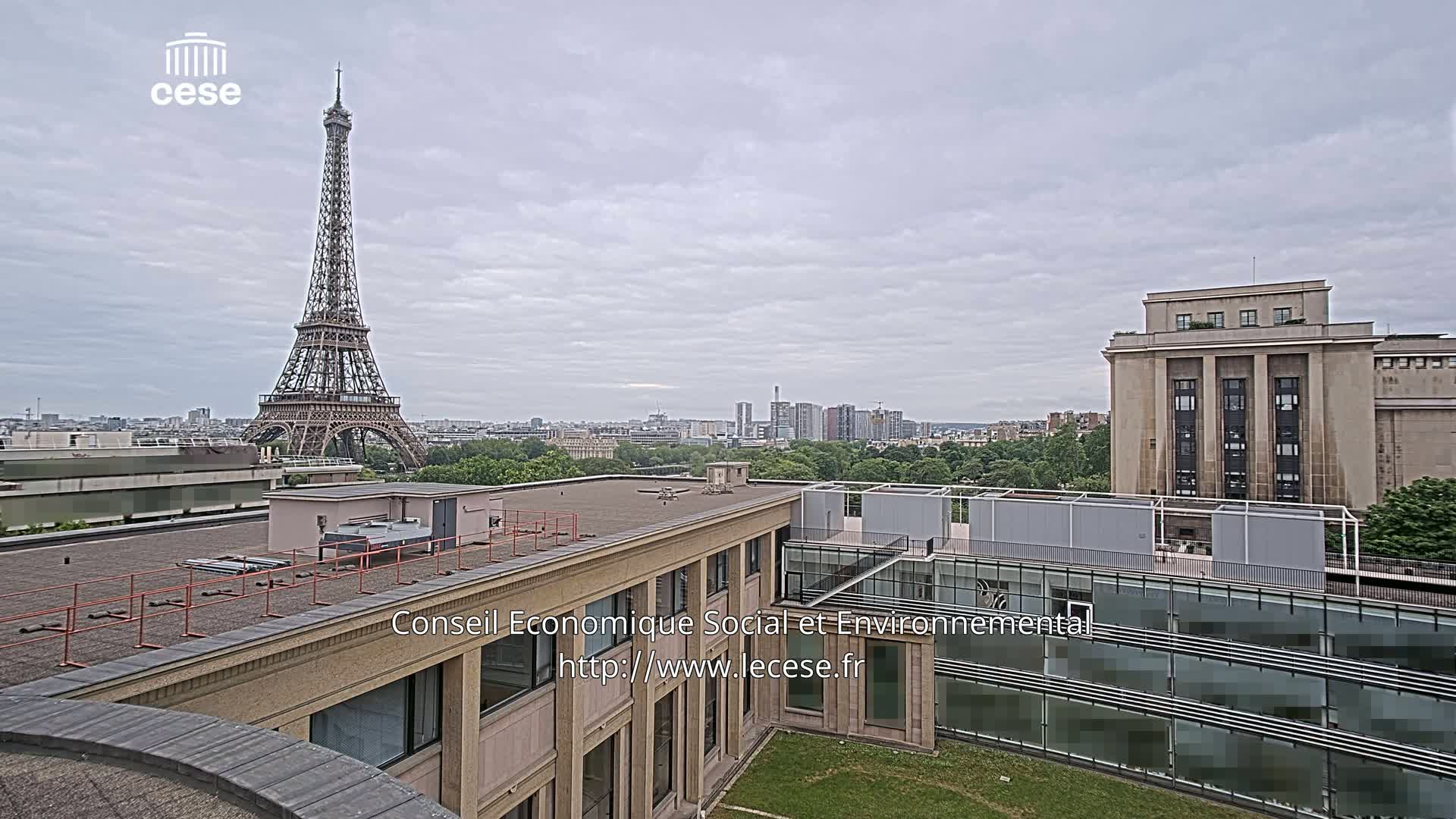 A view from a rooftop shows the Eiffel Tower in the distance under a cloudy sky, with modern and classical Parisian buildings in the foreground.