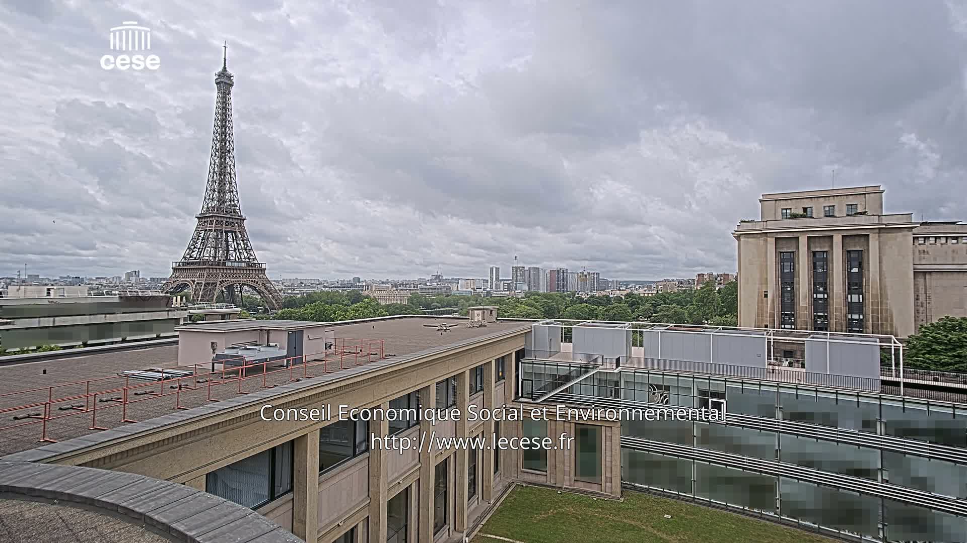 A view from a rooftop shows the Eiffel Tower in the distance under a cloudy sky, with other buildings and greenery in the foreground.