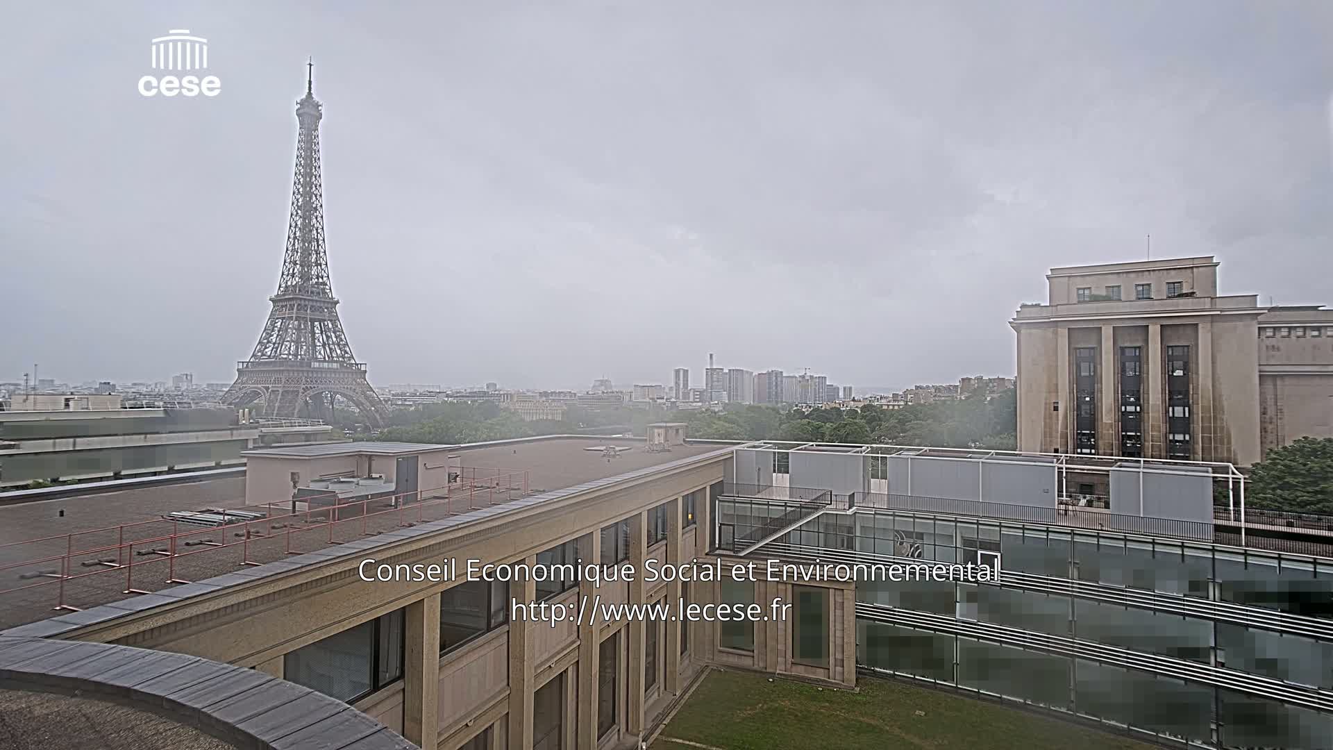 A view of the Eiffel Tower in the distance on an overcast day, from a rooftop overlooking a city with modern and classical buildings.