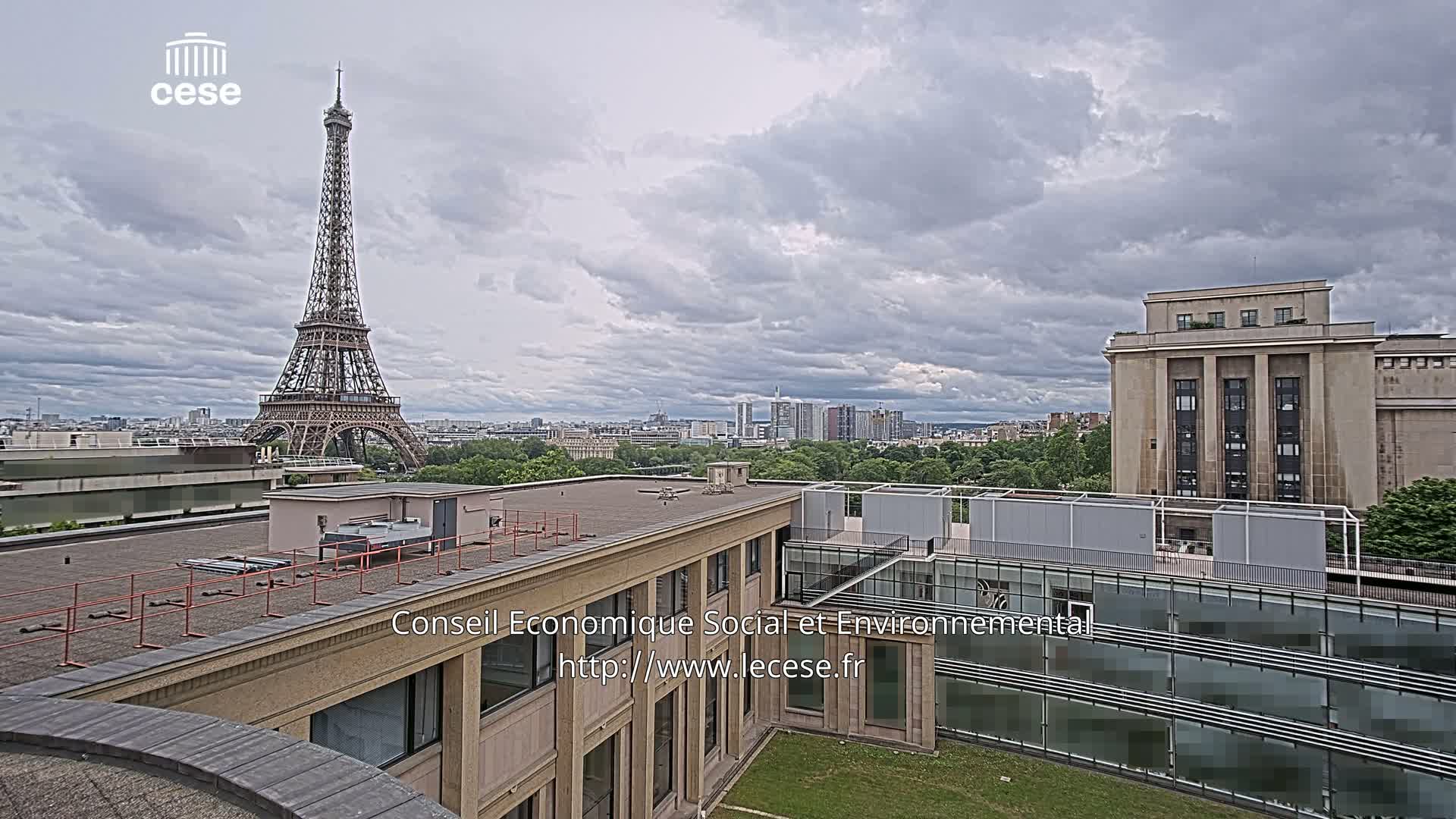 A view from a rooftop shows the Eiffel Tower in the distance under a cloudy sky, with a city skyline and other buildings visible.