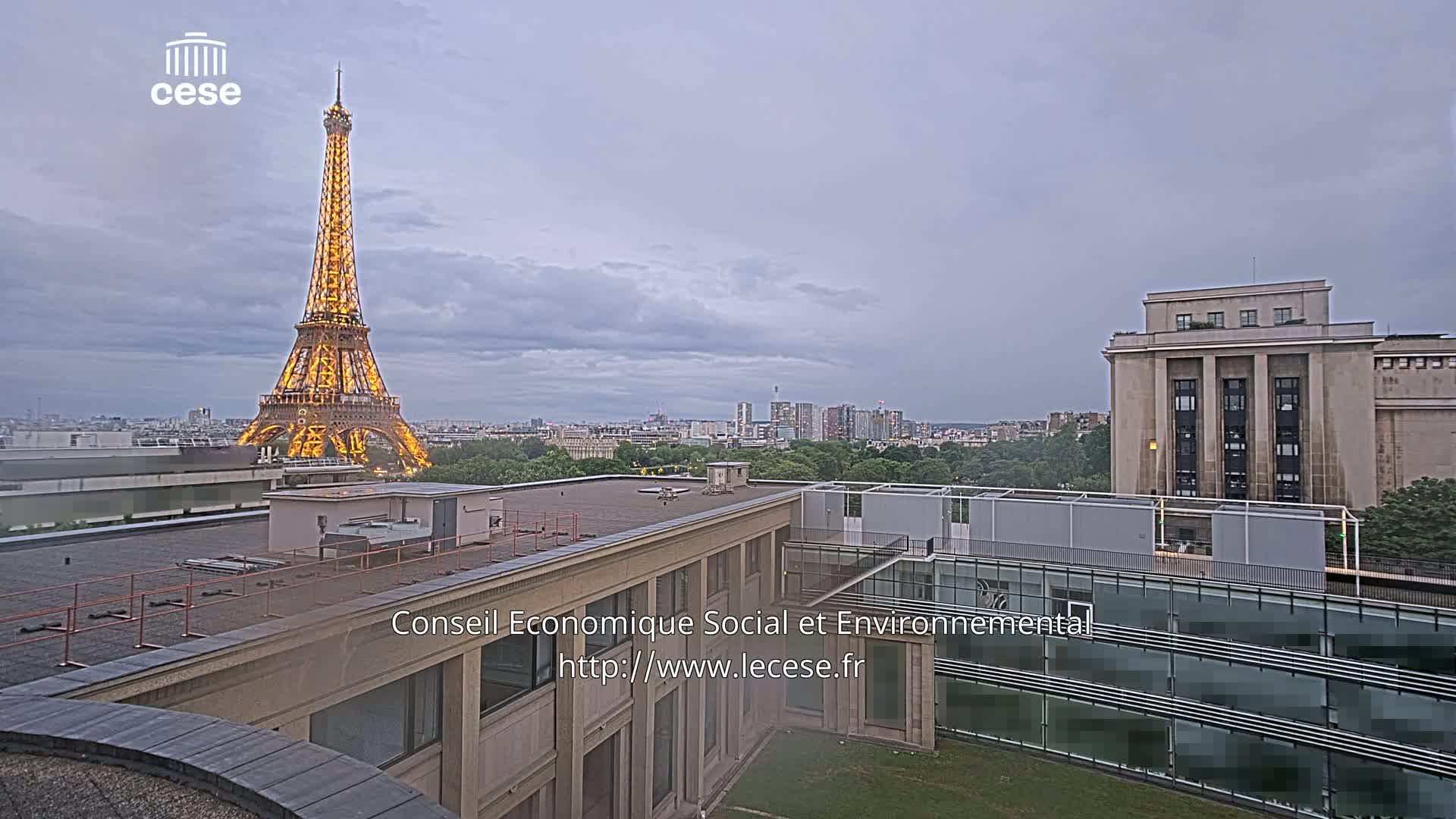 The Eiffel Tower is illuminated against an overcast sky, visible from a rooftop overlooking a city landscape.