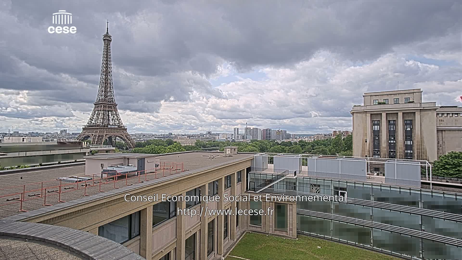 A view from a rooftop shows the Eiffel Tower in the distance under a cloudy sky, with a large building in the foreground and a cityscape beyond.