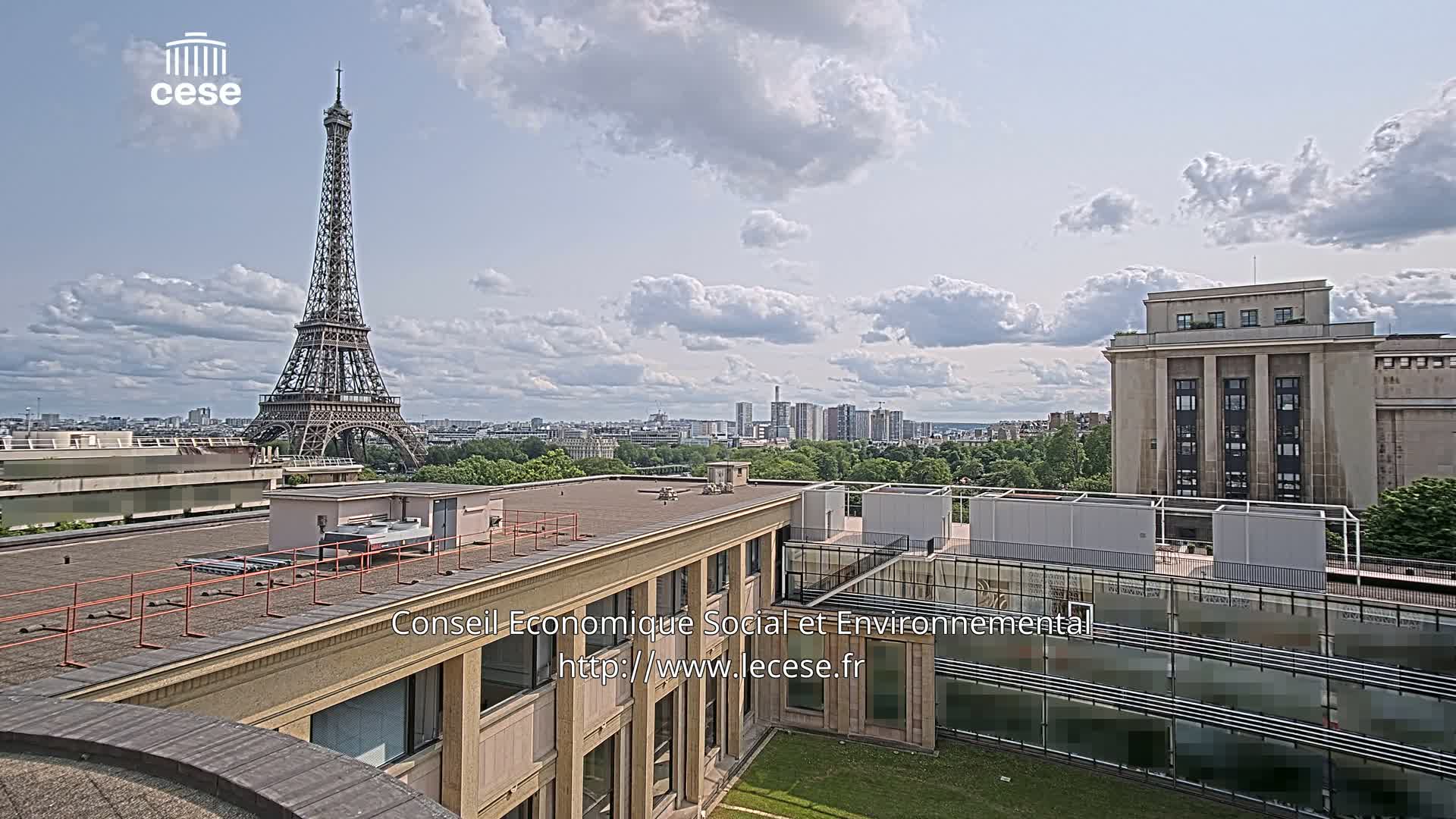 A rooftop view of Paris on a partly cloudy day shows the Eiffel Tower in the distance, along with other buildings and a cityscape.