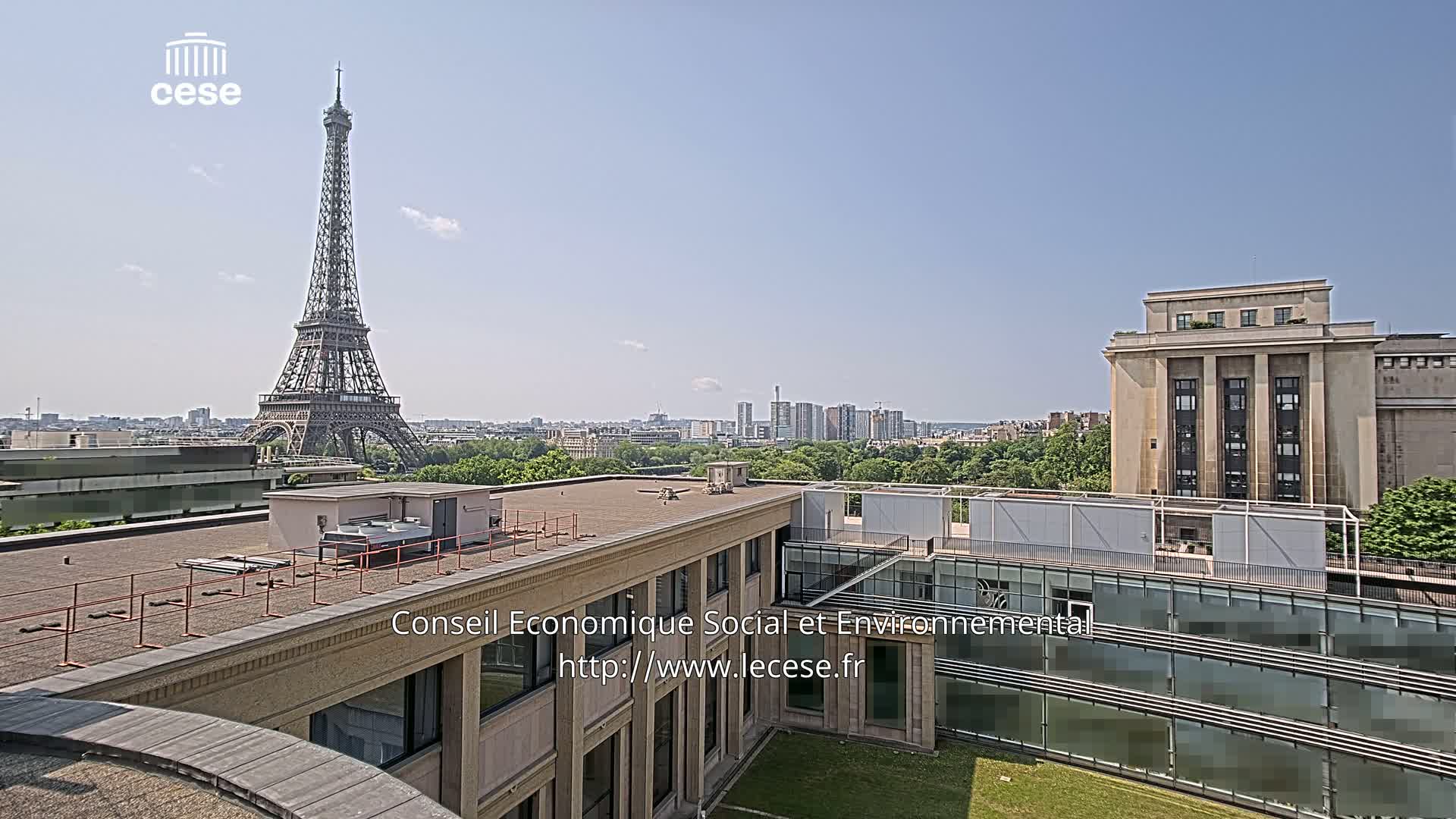 A rooftop view of Paris on a sunny day shows the Eiffel Tower in the distance, alongside city buildings and green spaces.