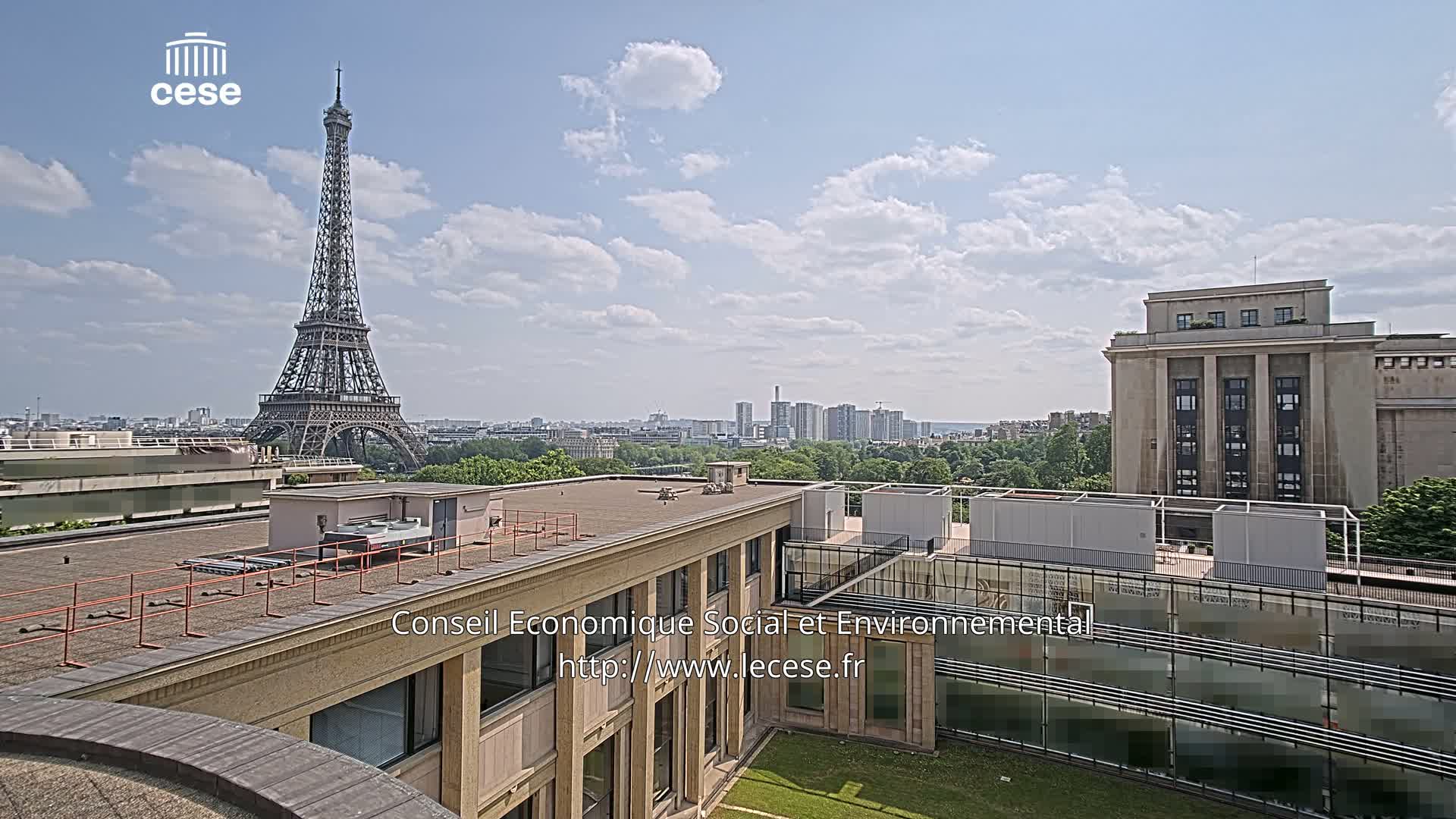 A rooftop view shows the Eiffel Tower in the distance under a partly cloudy sky, with a large building and cityscape in the background.