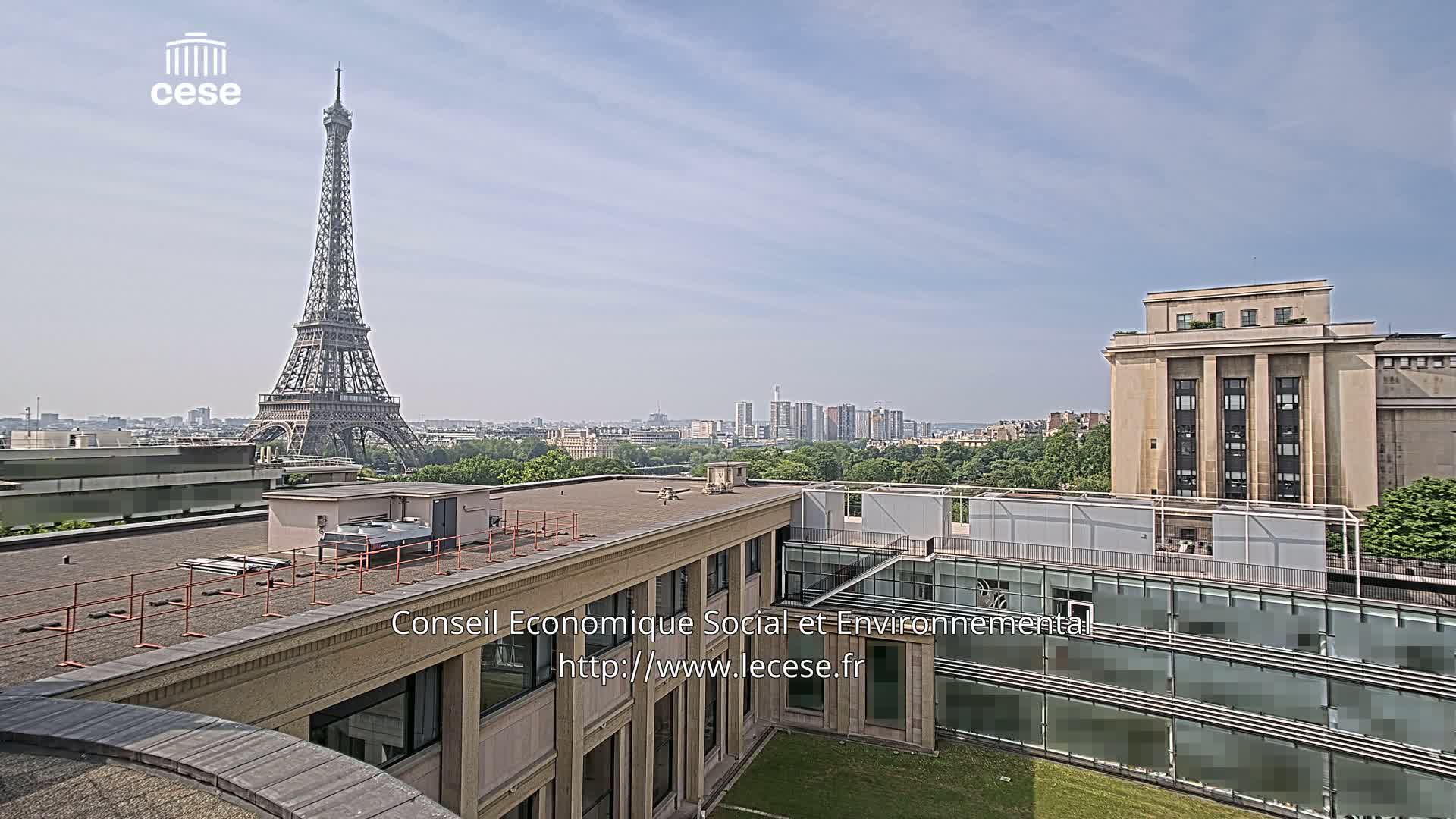A rooftop view of Paris on a partly sunny day shows the Eiffel Tower in the distance, alongside modern and classical buildings.