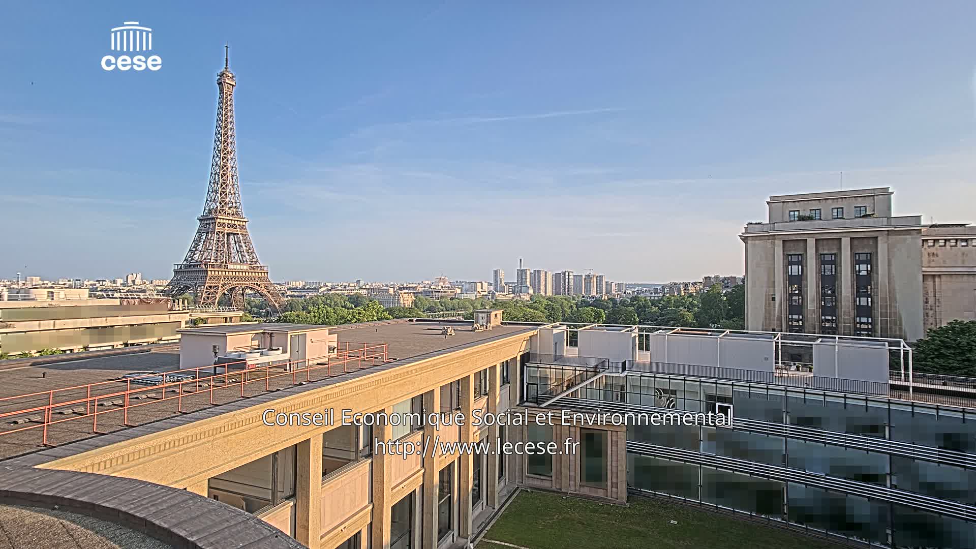 A rooftop view of Paris on a clear, sunny day shows the Eiffel Tower in the distance and a city skyline beyond a tree-lined area.