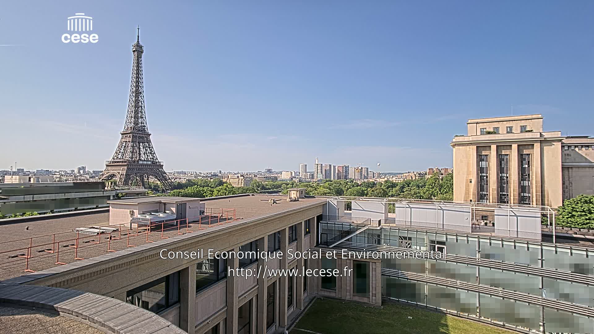 A rooftop view shows the Eiffel Tower in the distance on a clear, sunny day, with other buildings and green spaces visible in the foreground and mid-ground.