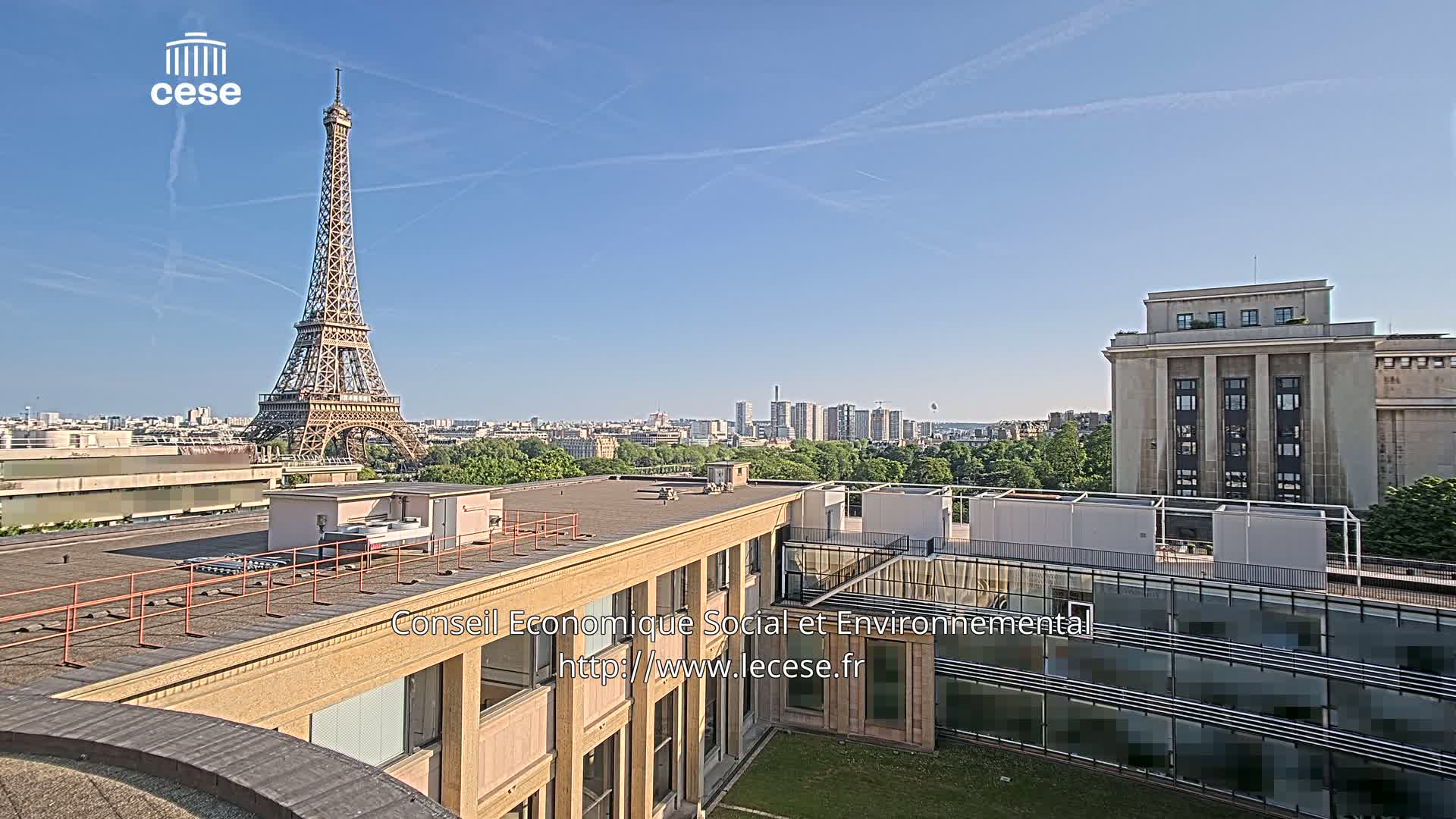 A rooftop view of Paris on a clear, sunny day shows the Eiffel Tower in the distance and a cityscape beyond a building's courtyard.