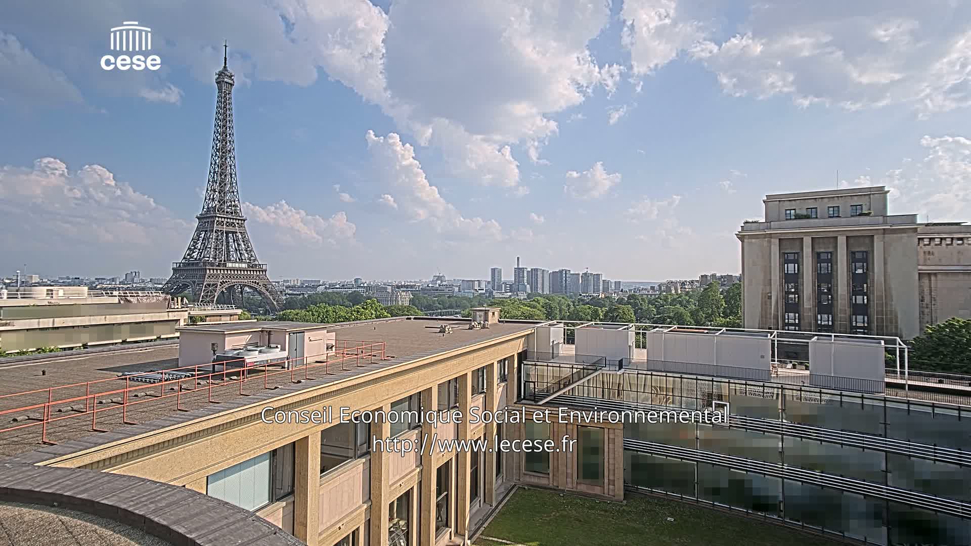 A rooftop view of Paris on a partly sunny day shows the Eiffel Tower in the distance and a large building in the foreground, with a city skyline visible beyond.