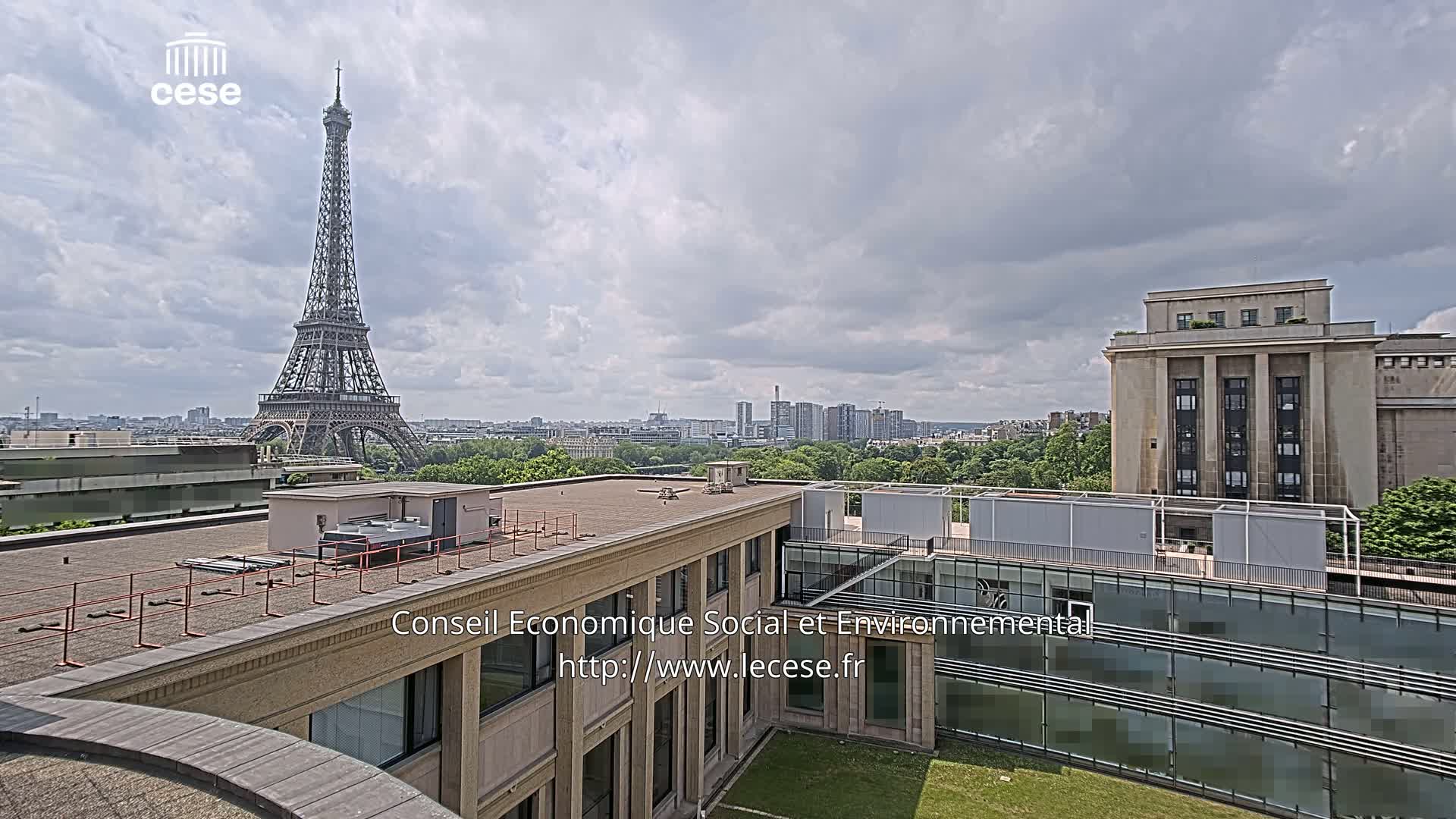 An elevated view shows the Eiffel Tower in the distance under a partly cloudy sky, with a city skyline and building rooftops in the foreground.
