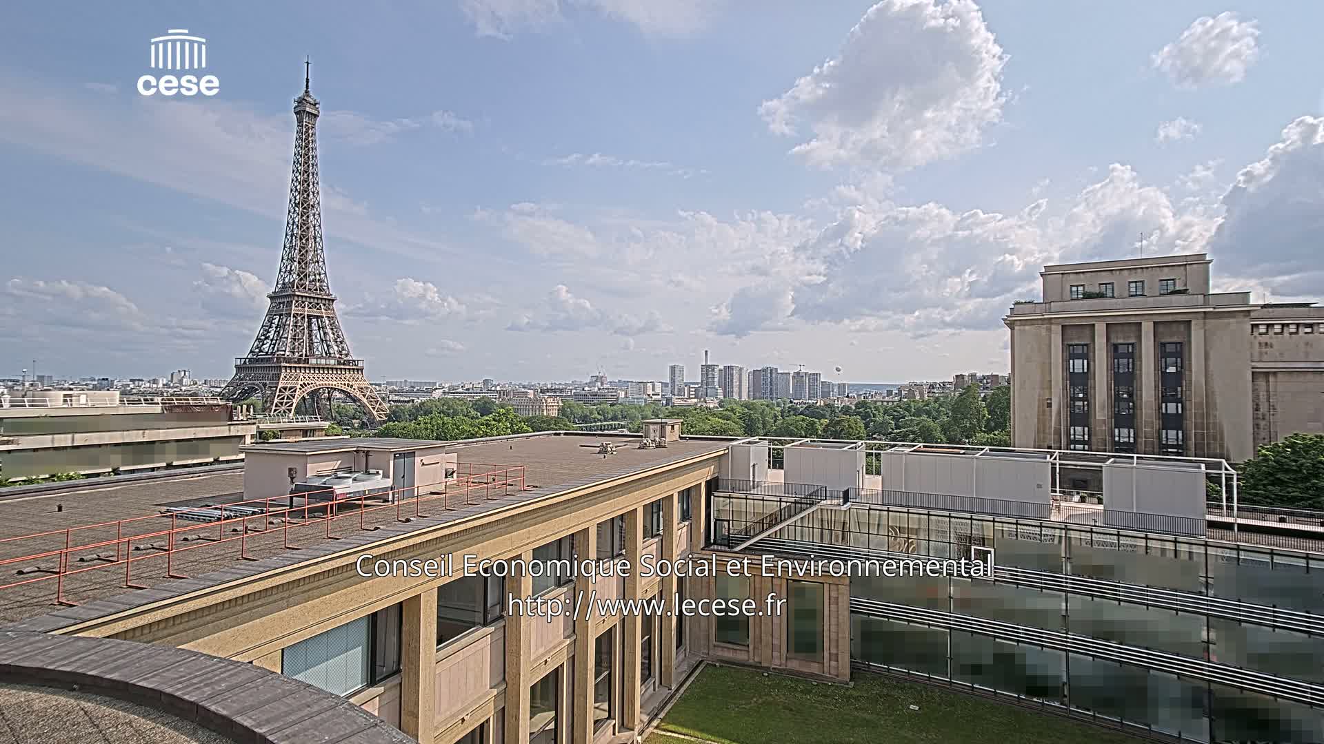 A rooftop view of the Eiffel Tower on a partly cloudy day, overlooking a city skyline and a large building with a flat roof.