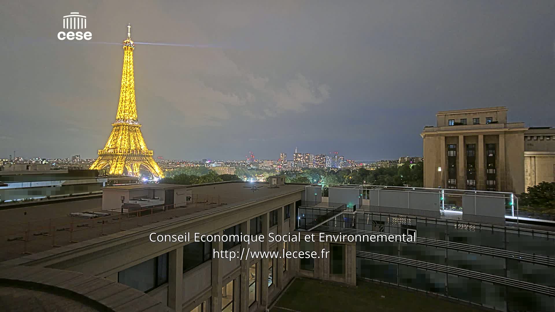 A rooftop view of the Eiffel Tower on a partly cloudy day, overlooking a city skyline and a large building with a flat roof.