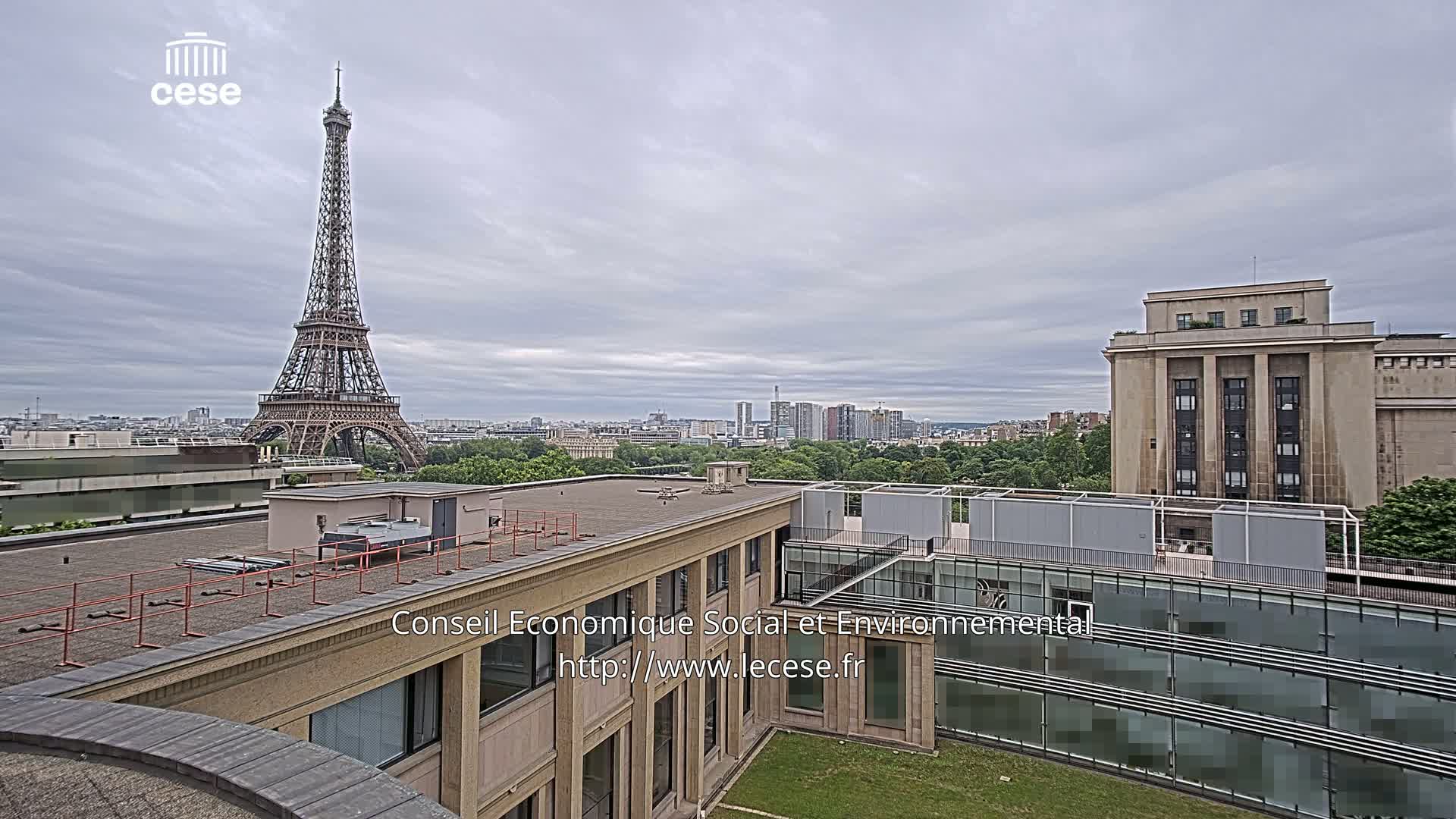 A view from a rooftop shows the Eiffel Tower in the distance under a cloudy sky, with a city skyline and other buildings nearby.
