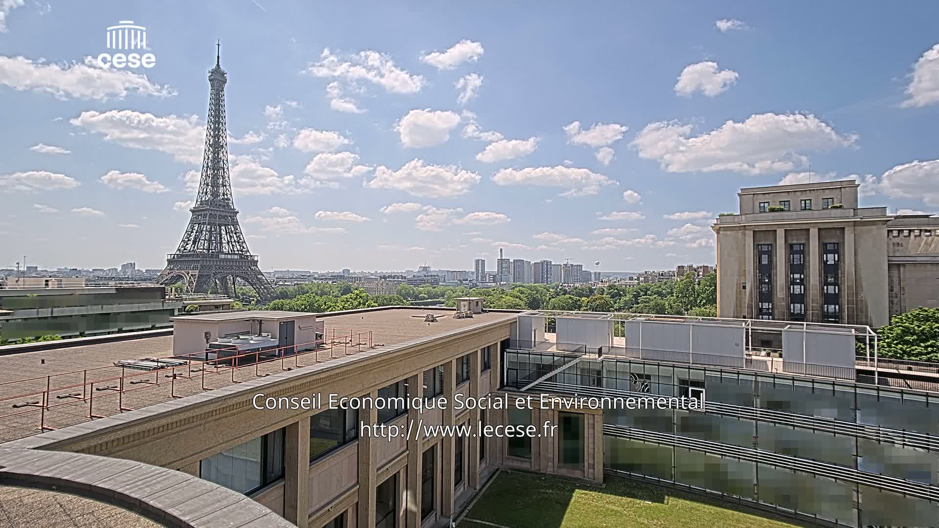 A rooftop view of Paris on a partly sunny day shows the Eiffel Tower in the distance and a large building in the foreground with a grassy courtyard.