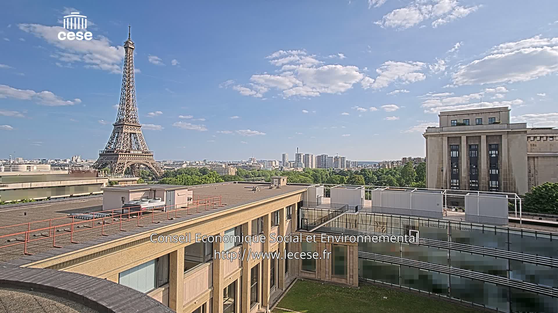 A rooftop view shows the Eiffel Tower in the distance under a partly cloudy sky, with a city skyline and another building nearby.