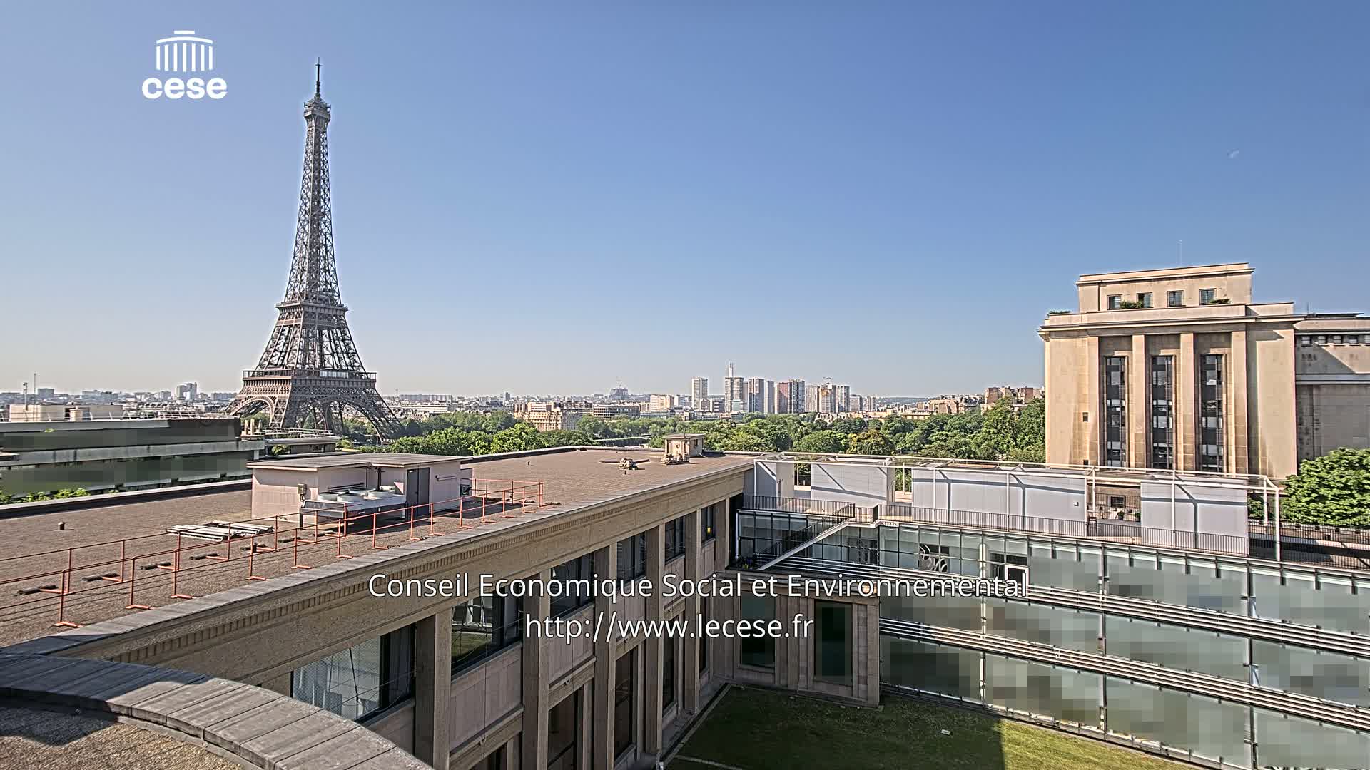 A rooftop view of Paris on a clear, sunny day shows the Eiffel Tower in the distance, with city buildings and greenery in the foreground.
