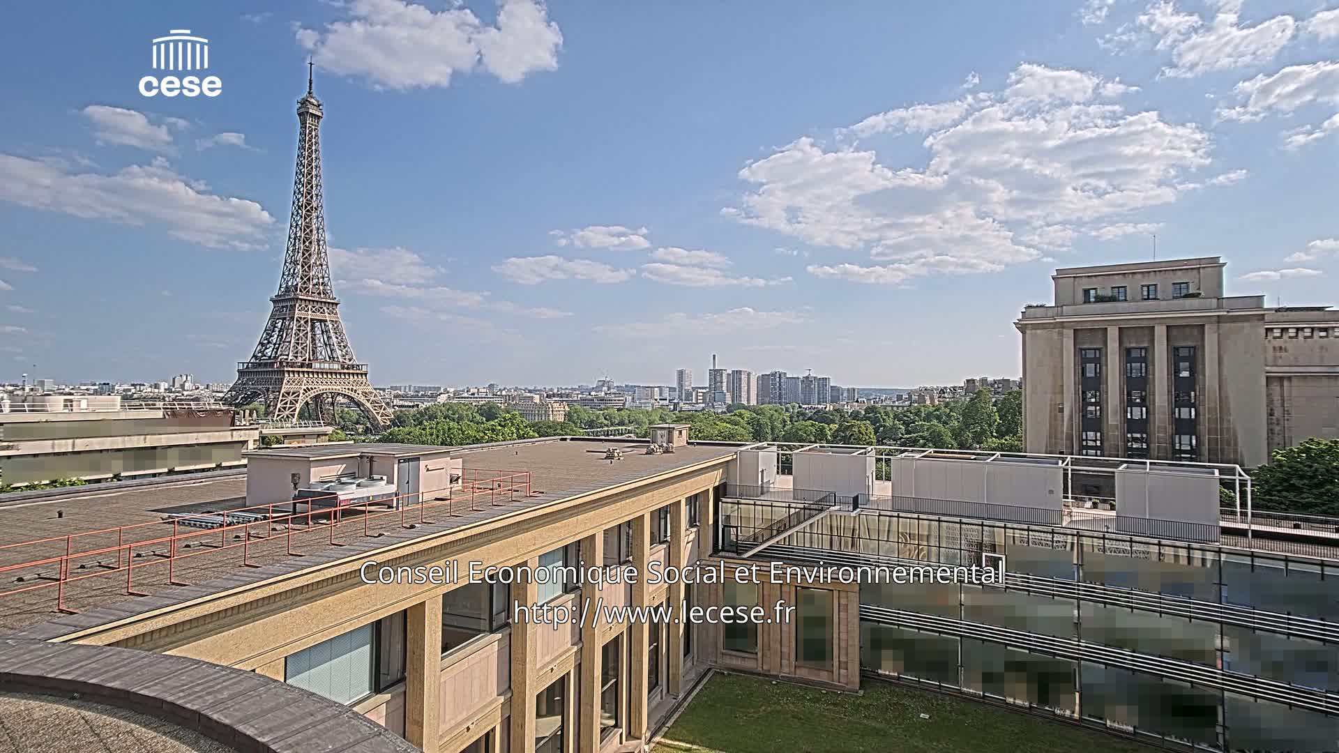 A rooftop view of Paris on a partly cloudy day shows the Eiffel Tower in the distance, with other buildings and greenery in the foreground.