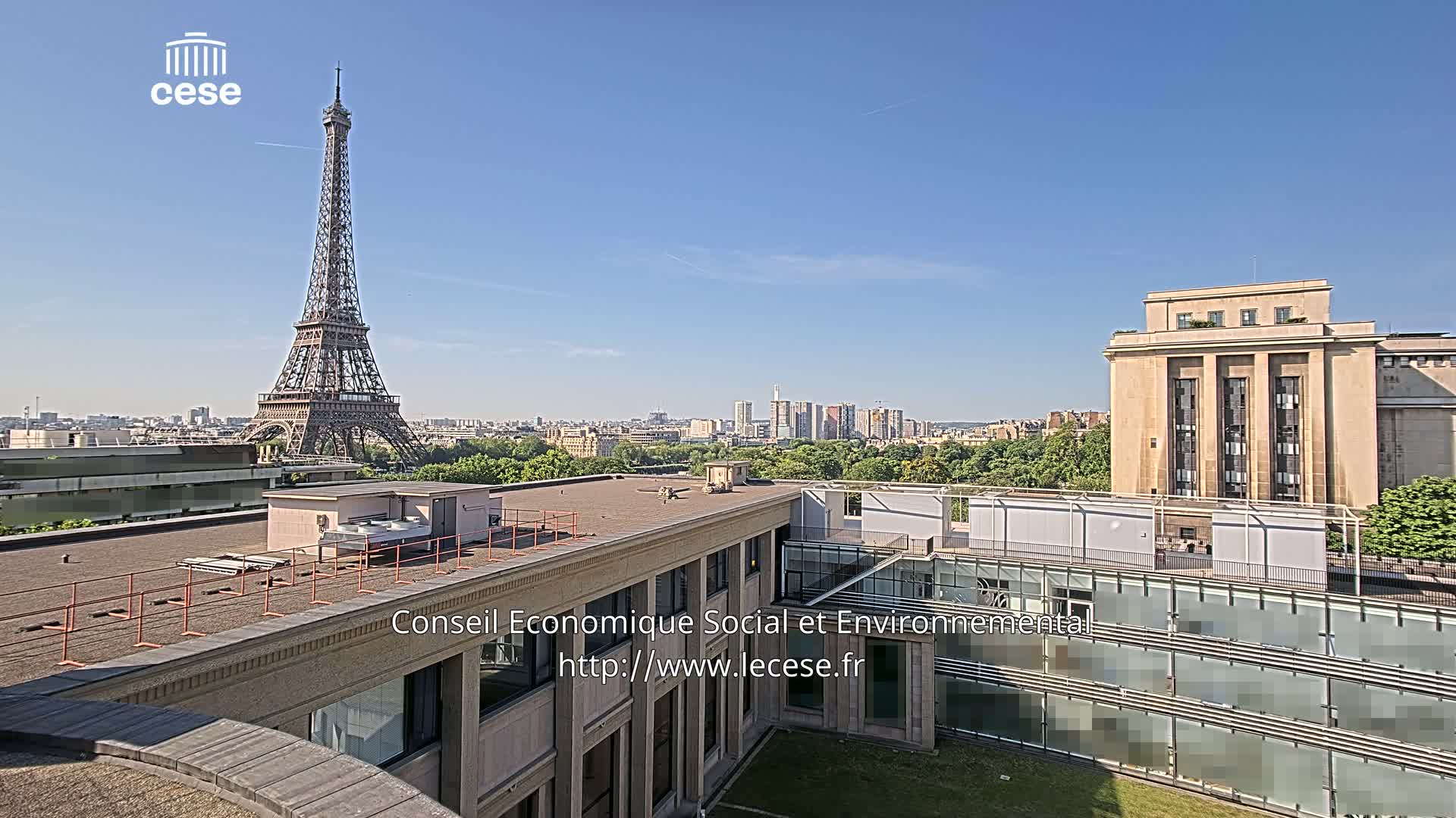 A rooftop view of Paris on a clear, sunny day shows the Eiffel Tower in the distance, city buildings, and a large building in the foreground.
