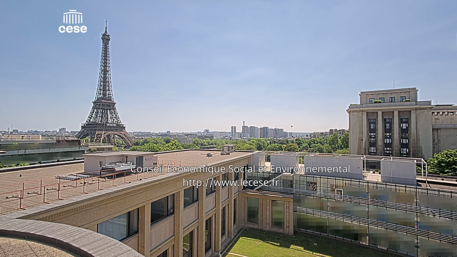A rooftop view shows the Eiffel Tower in the distance on a clear, sunny day, with a large building in the foreground and a cityscape in between.