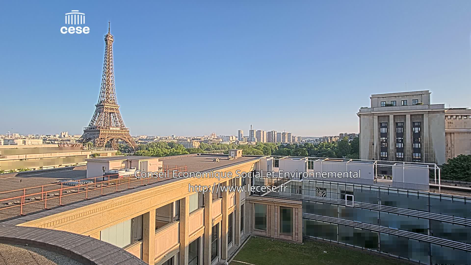 A view from a rooftop shows the Eiffel Tower in the distance on a clear, sunny day, with other city buildings and greenery in the foreground.