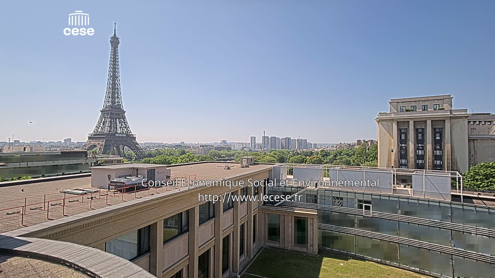 A rooftop view of a city on a sunny day, featuring the Eiffel Tower in the distance and a modern building in the foreground.