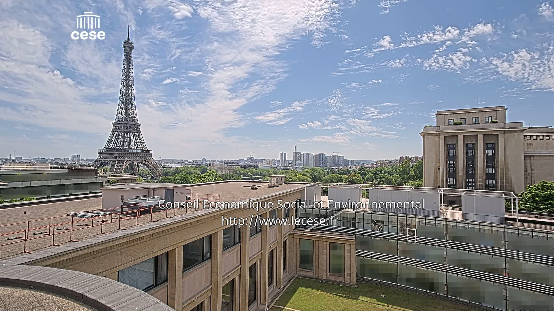 A view from a rooftop shows the Eiffel Tower in the distance under a partly cloudy sky, with a city skyline and a large building in the foreground.