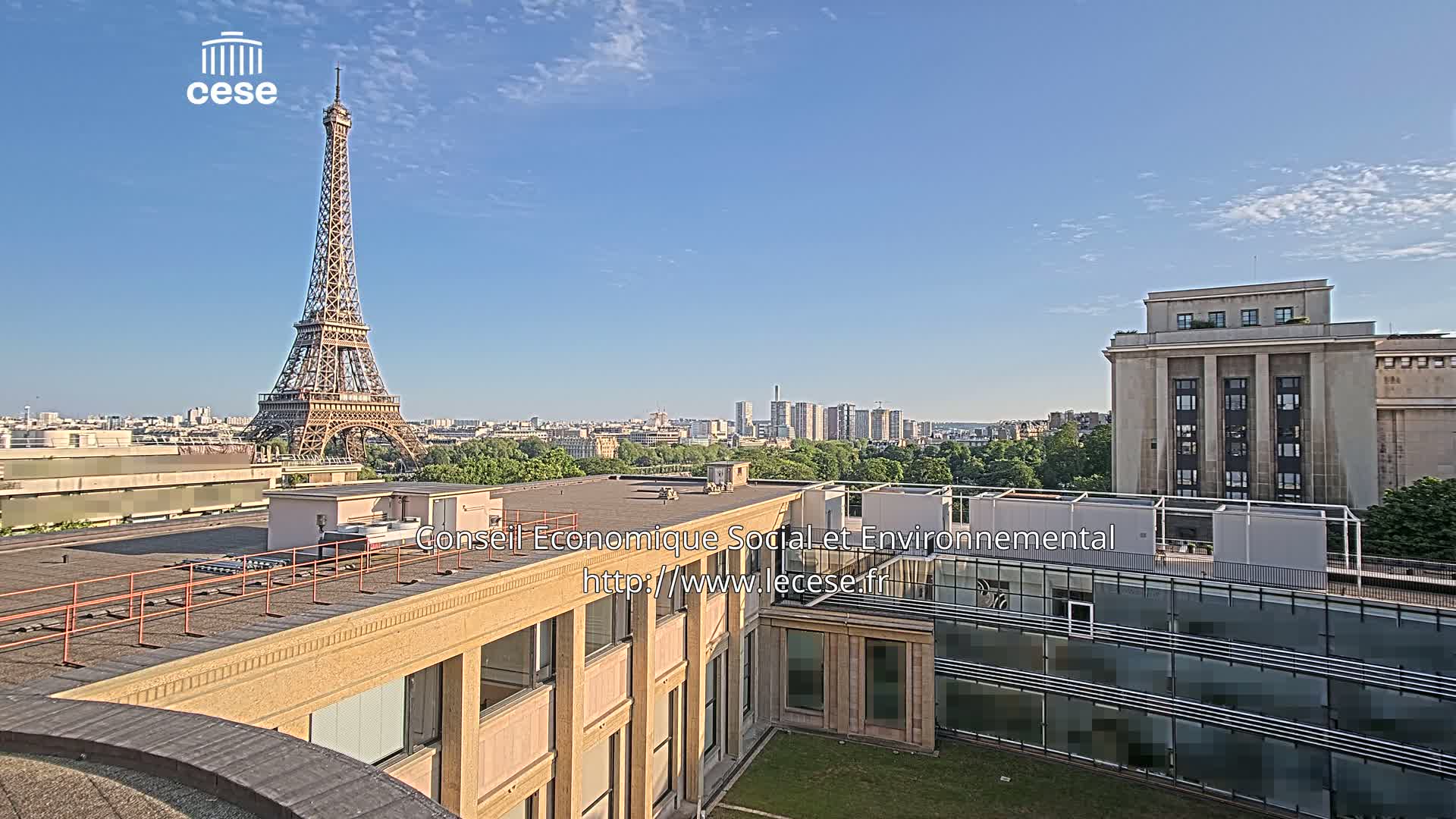 A rooftop view of the Eiffel Tower in the distance on a clear, sunny day, with a large building and cityscape visible in the foreground.
