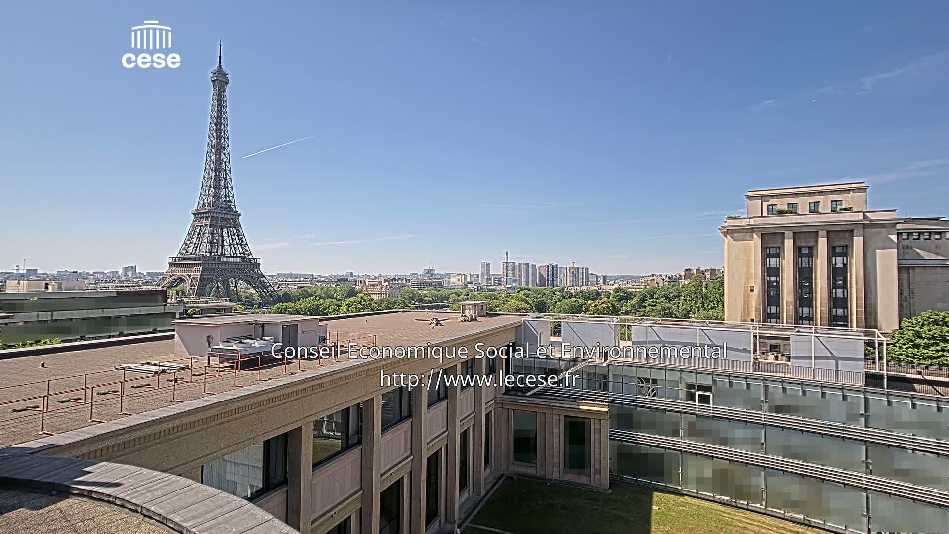 A rooftop view shows the Eiffel Tower in the distance under a clear blue sky, overlooking a cityscape and adjacent buildings.