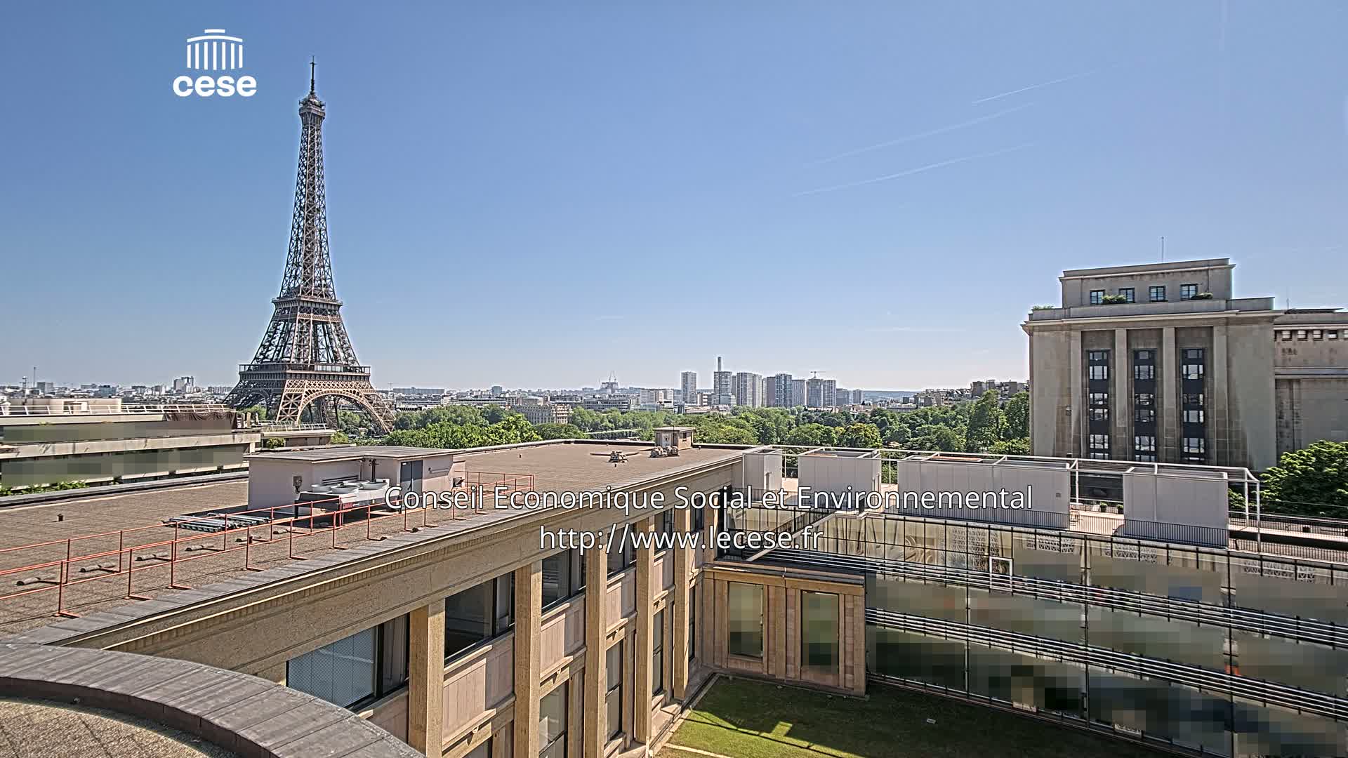 A rooftop view of Paris on a clear, sunny day shows the Eiffel Tower in the distance and a large building in the foreground.