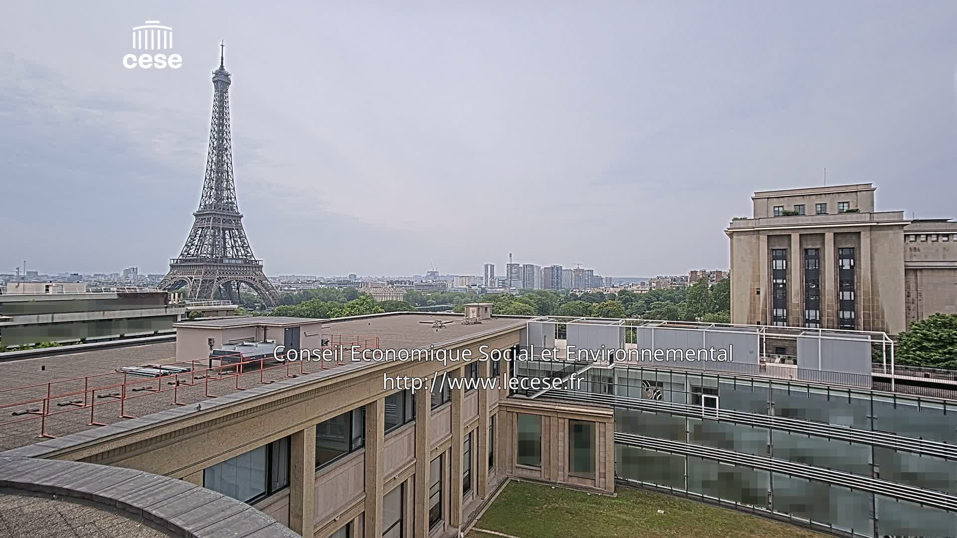 A rooftop view shows the Eiffel Tower in the distance under an overcast sky, with other buildings and greenery in the foreground.