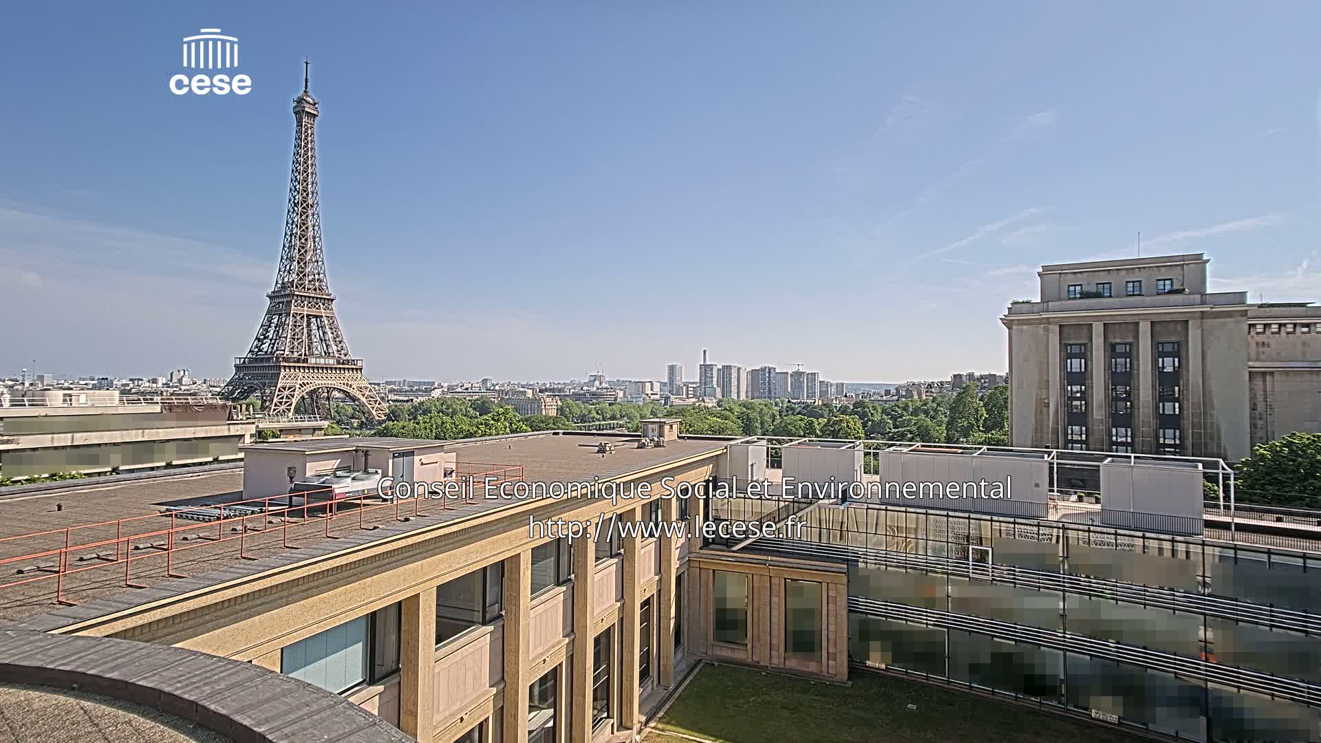 A rooftop view of a city on a clear, sunny day, featuring the Eiffel Tower in the distance and a large building in the foreground.