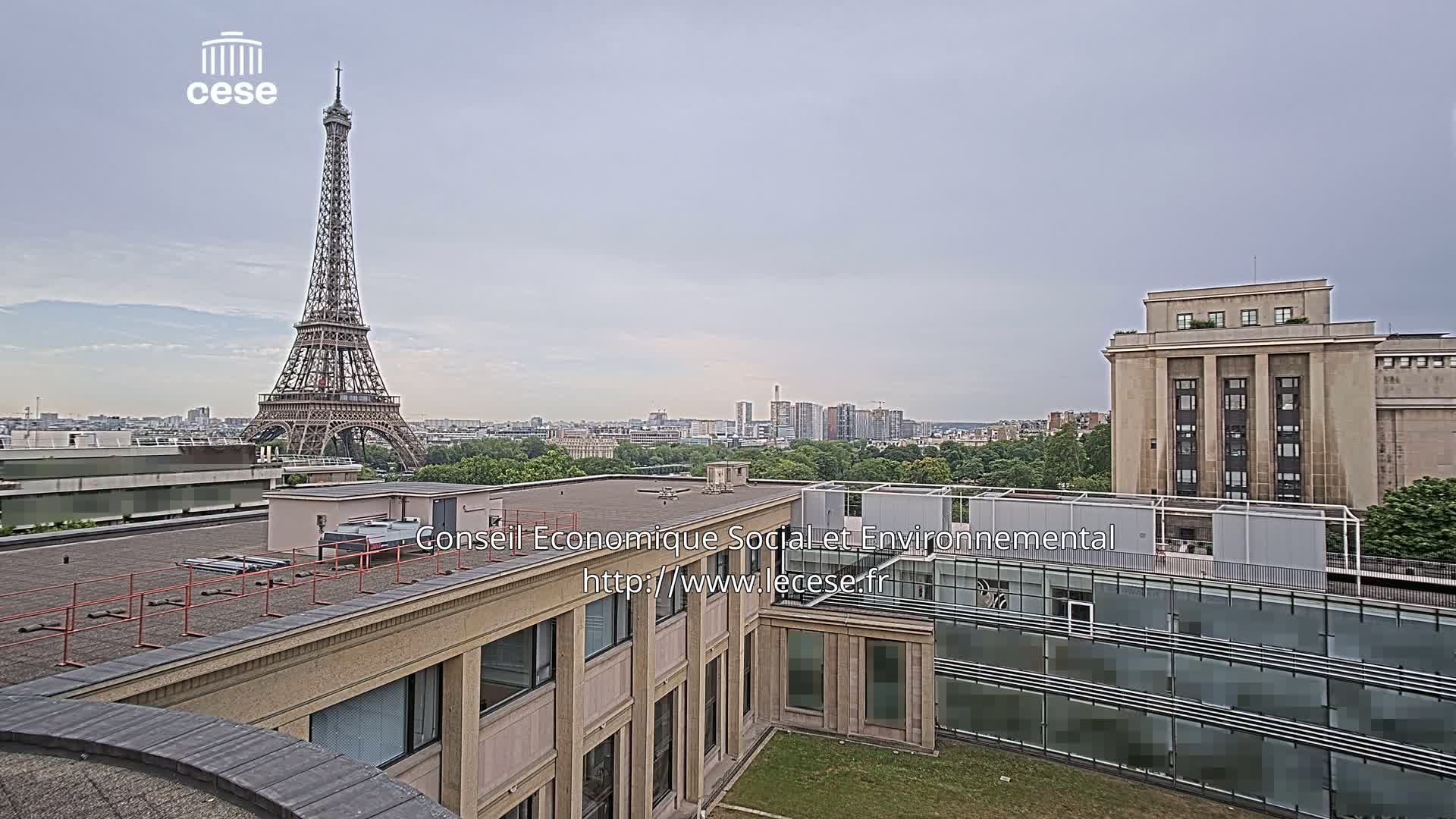 A view from a rooftop shows the Eiffel Tower in the distance under an overcast sky, with other buildings and green spaces in the foreground.