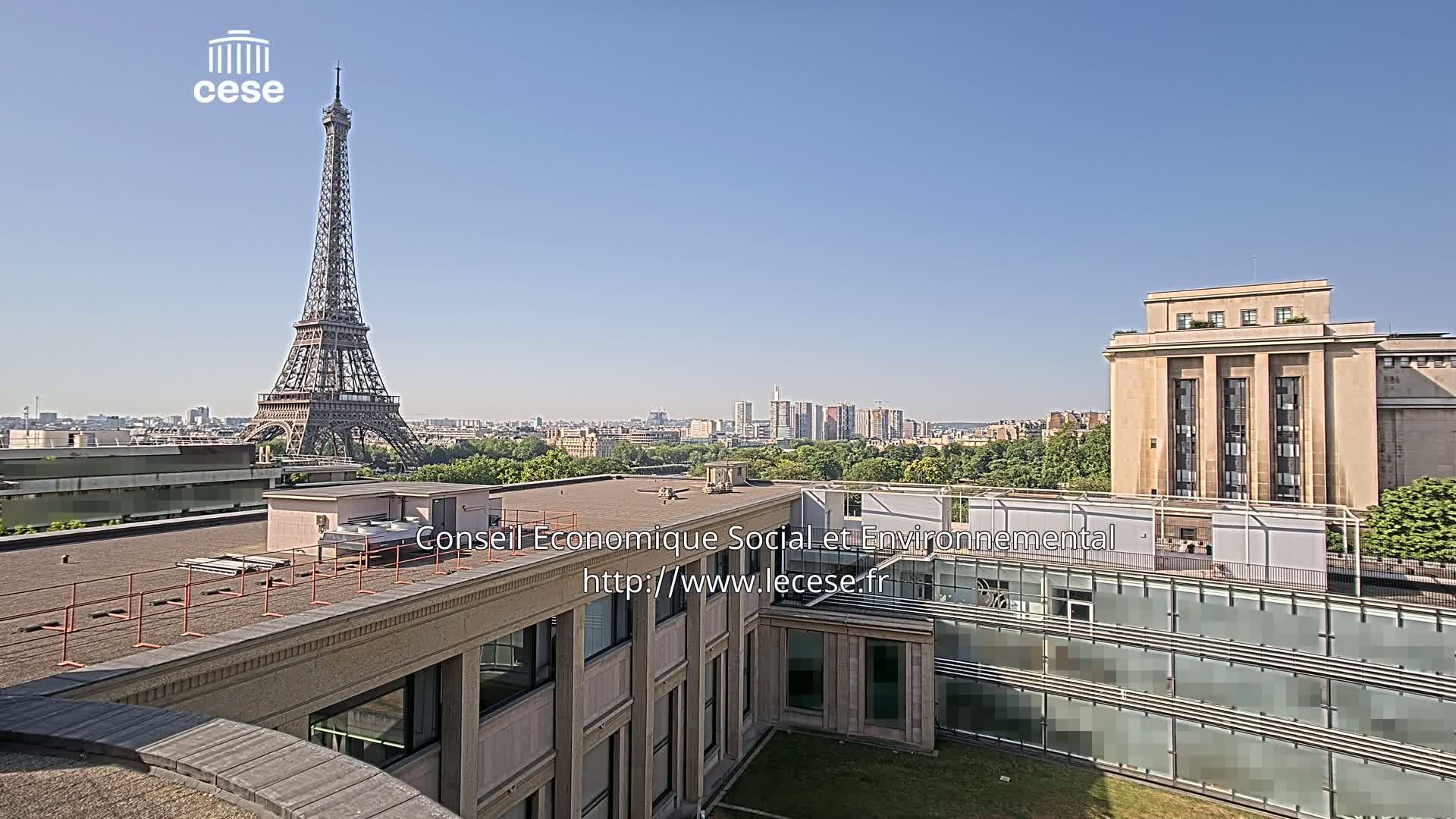 A rooftop view shows the Eiffel Tower in the distance under a clear blue sky, with a large building in the foreground and other buildings and trees extending to the horizon.