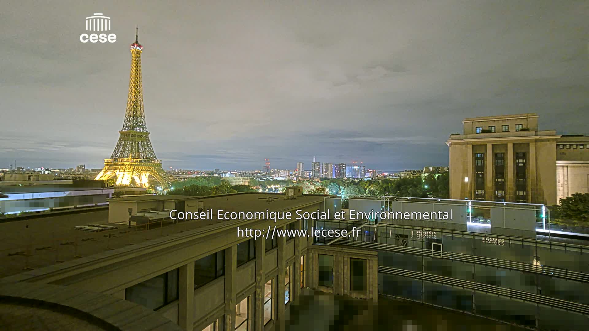 A rooftop view shows the Eiffel Tower in the distance under a cloudy sky, with other buildings and trees in the foreground.