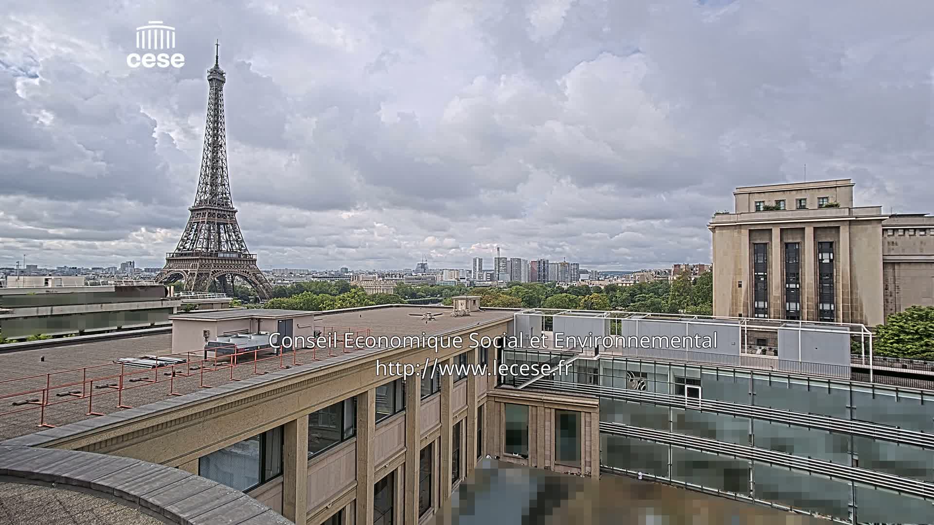 An outdoor view of the Eiffel Tower in the distance under a cloudy sky, with a modern building and cityscape in the foreground.