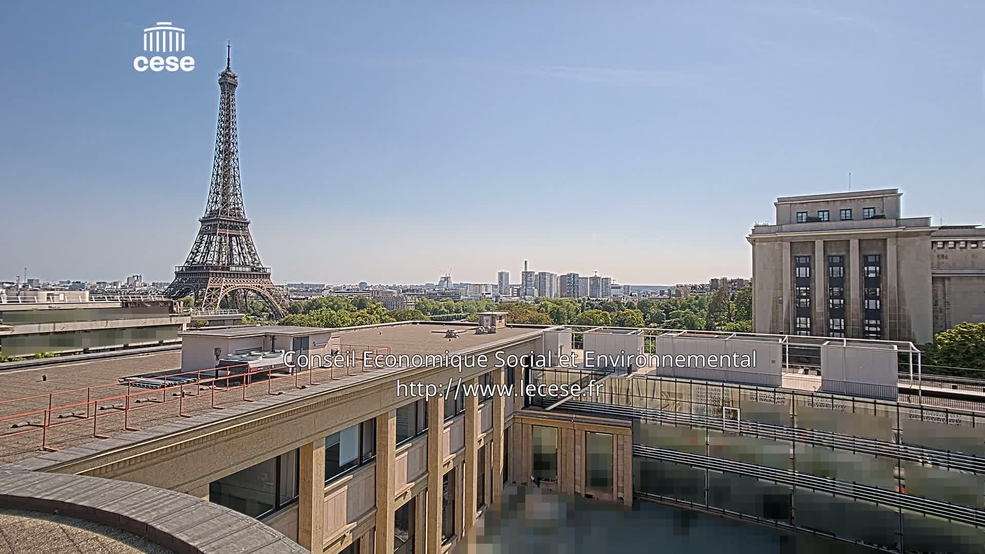 An outdoor view of the Eiffel Tower in the distance under a cloudy sky, with a modern building and cityscape in the foreground.