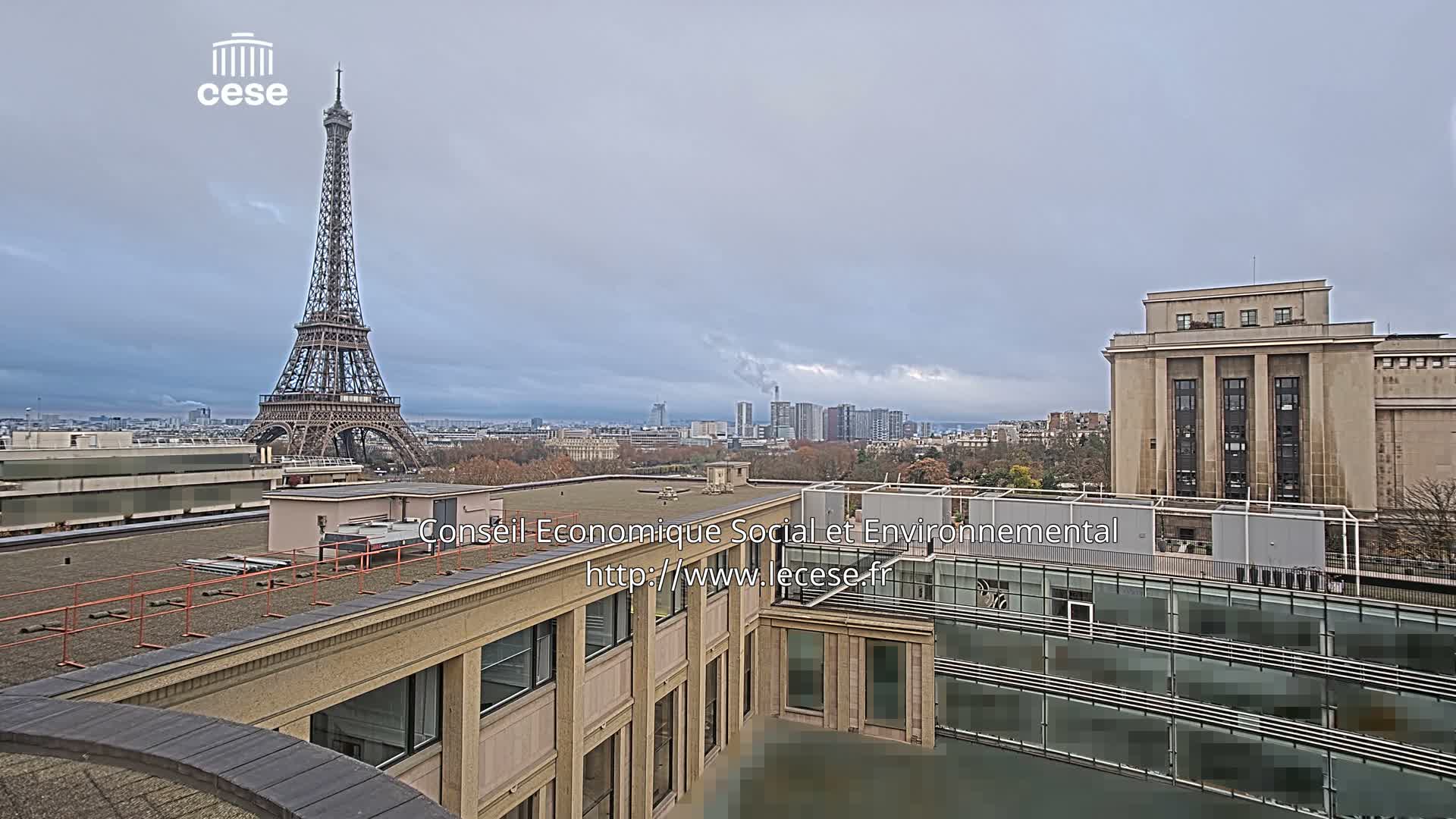 A cloudy, grey sky hangs over a Parisian cityscape featuring the Eiffel Tower prominently on the left, with various buildings in the midground and foreground.