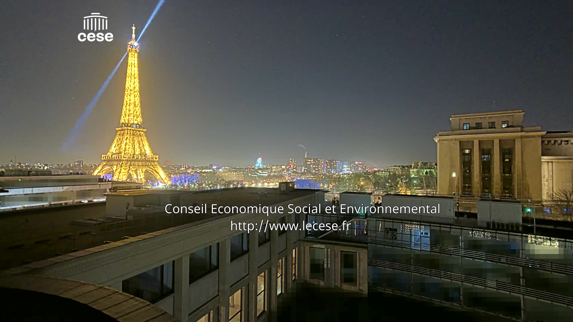 A clear night in Paris reveals the brightly lit Eiffel Tower beaming light into a star-dotted sky, surrounded by other illuminated city buildings and foreground rooftops.