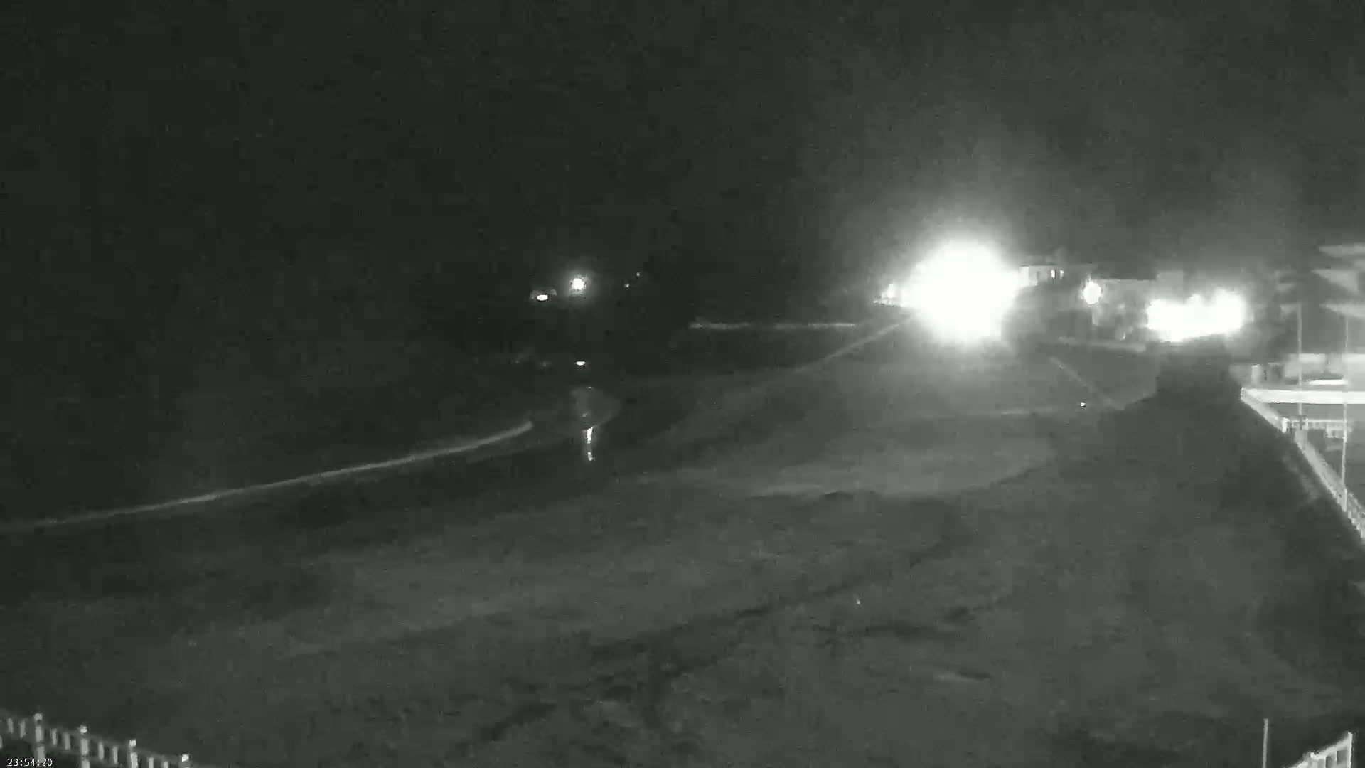 A nighttime, low-light view of a dark beach with a road and buildings in the background, possibly under cloudy conditions.