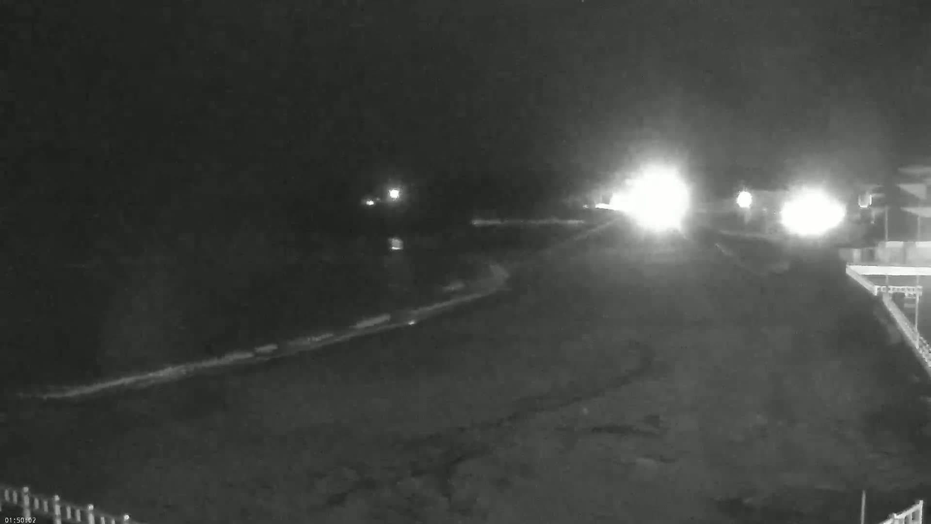 A nighttime, low-light view shows a dark beach with a gently curving shoreline and vehicle headlights in the distance.