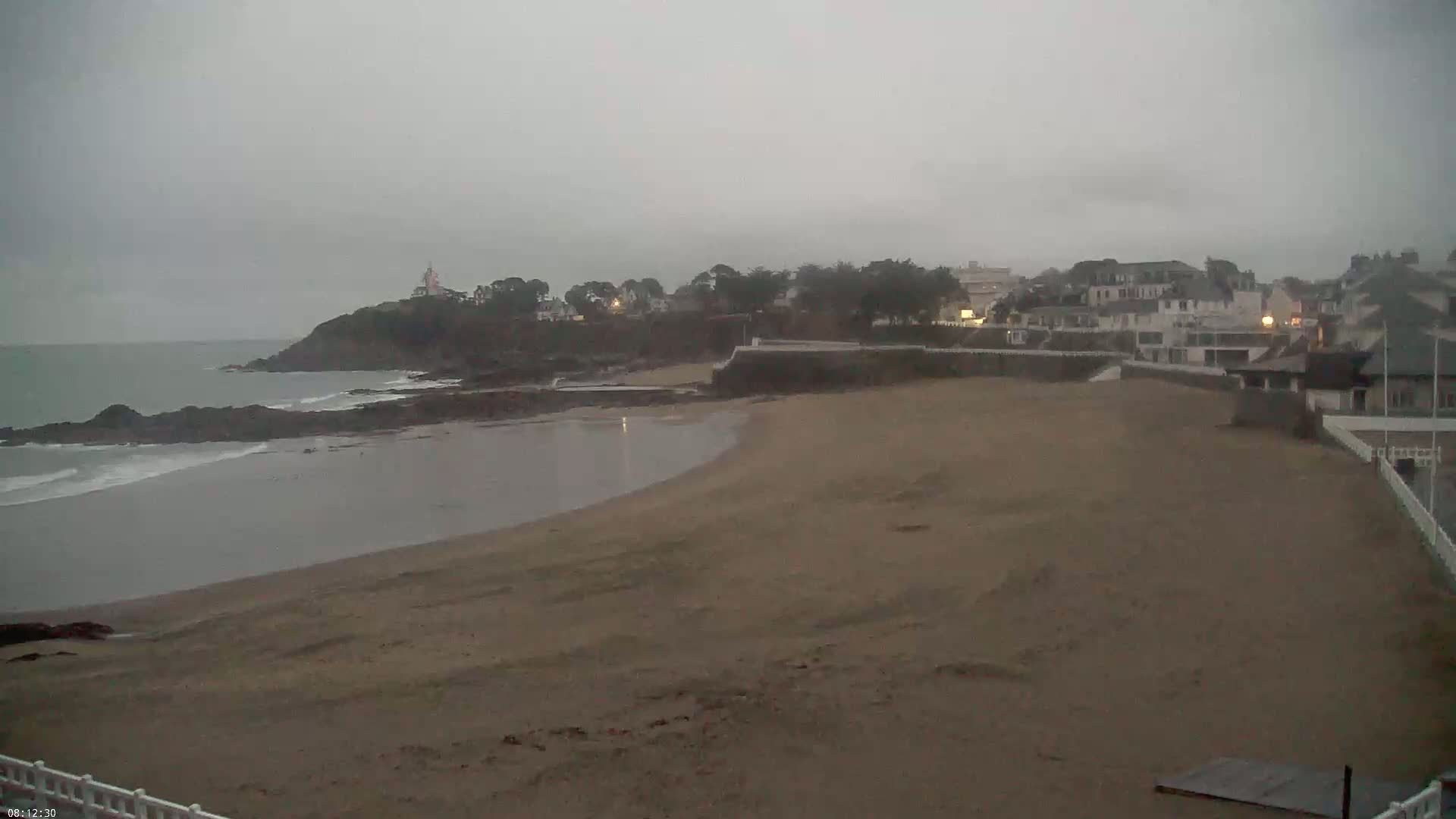 A sandy beach curves into a calm bay under an overcast sky, with a small town and a lighthouse visible in the distance.