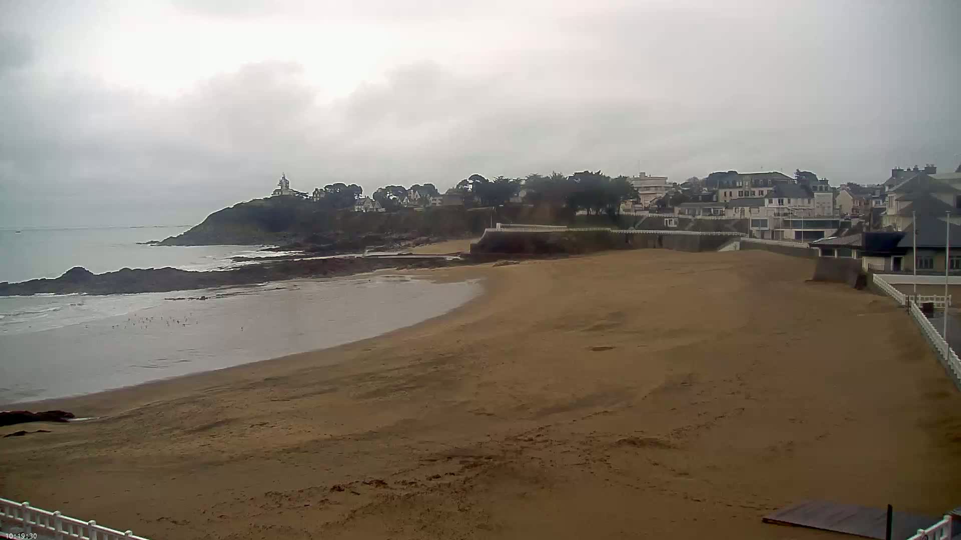 A sandy beach curves around a rocky shoreline, under an overcast sky, with a small town and a structure on a hill in the background.