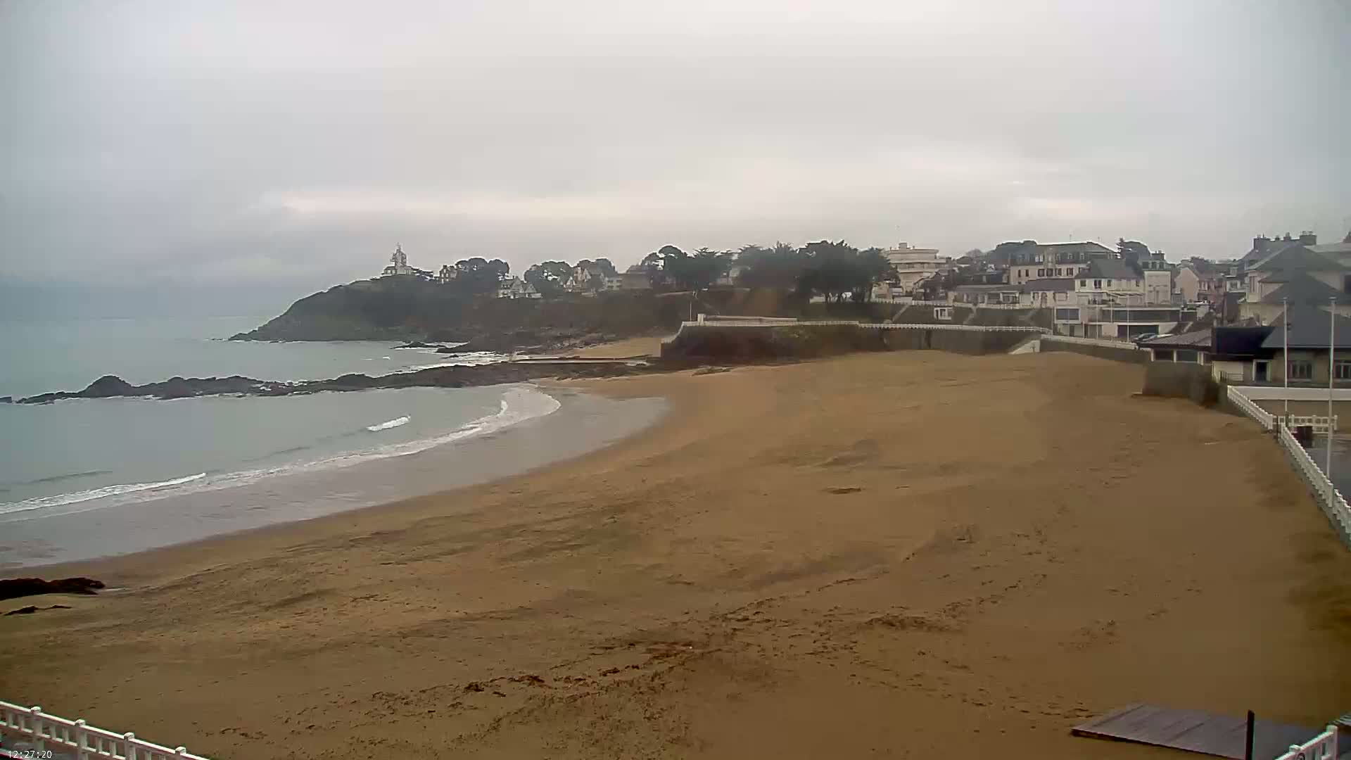 A sandy beach curves gently around a rocky point, meeting a calm, gray sea under an overcast sky, with buildings and trees visible on the cliffside.