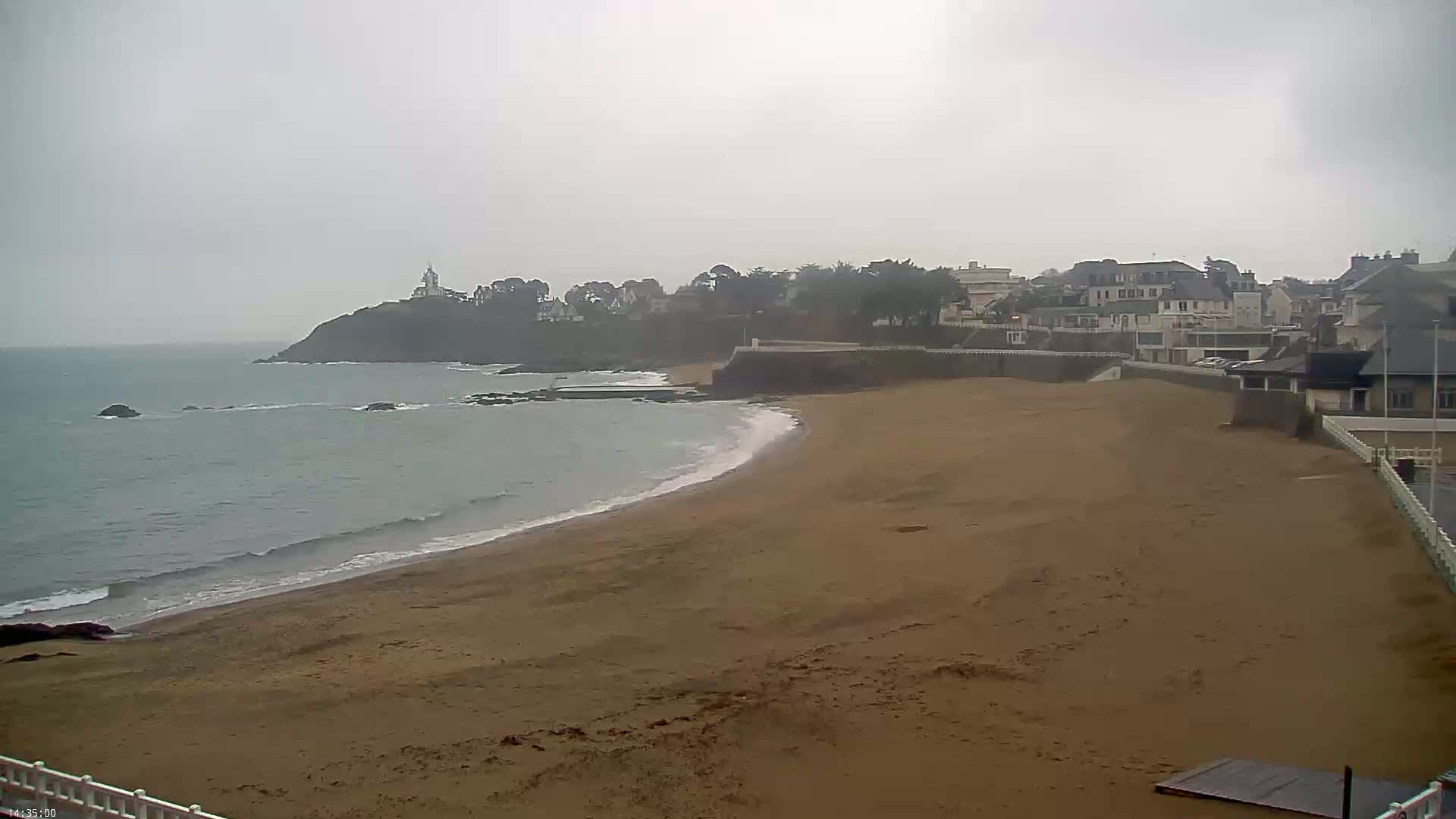 A sandy beach curves gently around a coastline, meeting a calm ocean under an overcast sky.
